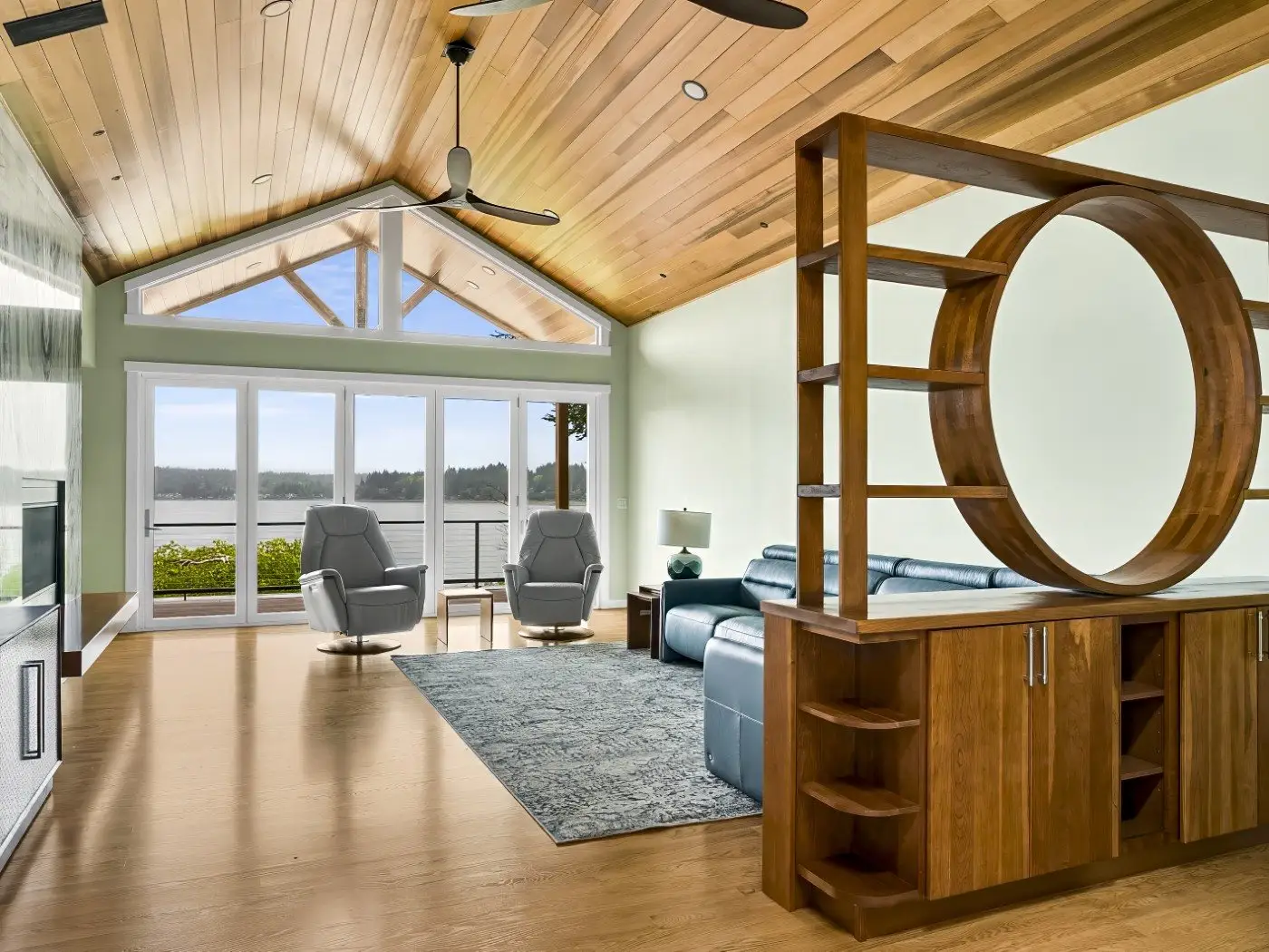 Modern living room with wood floors and ceiling, large windows overlooking water, blue sectional sofa, two gray chairs, a blue rug, and a unique wooden shelving unit with a circular design.