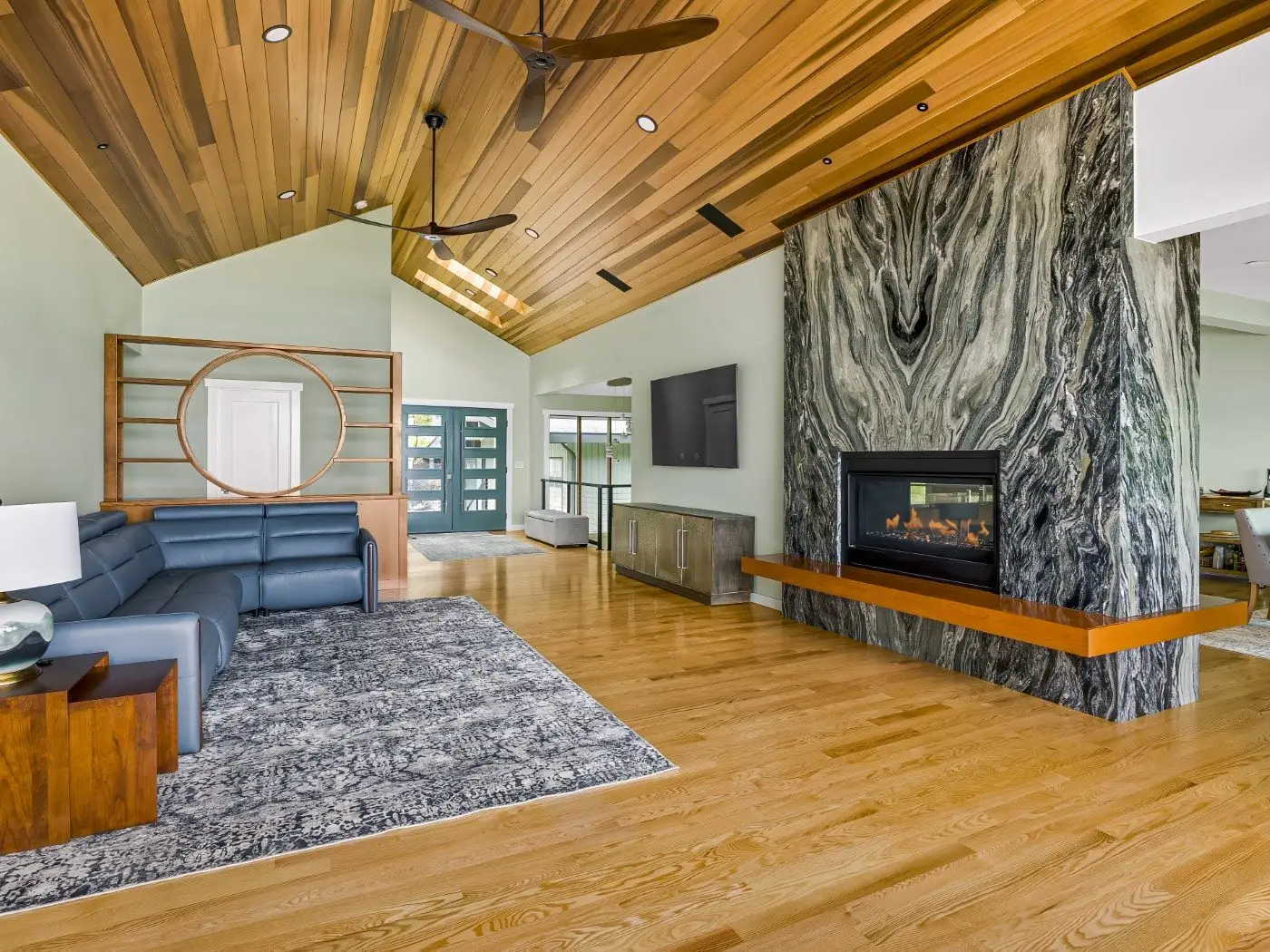 Modern living room with wood floors and ceiling, a large blue sectional sofa, patterned rug, marble fireplace with a wooden mantel, TV, and decorative shelving; bright with natural light.