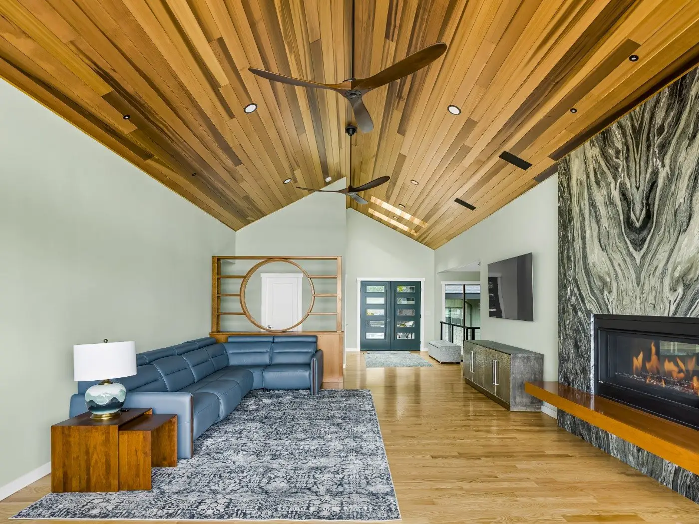 Modern living room with a wood-paneled vaulted ceiling, blue sectional sofa, large area rug, stone fireplace, built-in shelving, and a wall-mounted TV. Glass doors and windows let in natural light.