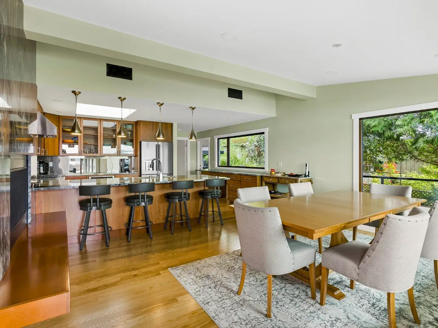 Modern open-concept kitchen and dining area with wood floors, a large island with barstools, pendant lights, wooden cabinets, a dining table with cushioned chairs, and large windows showing greenery outside.