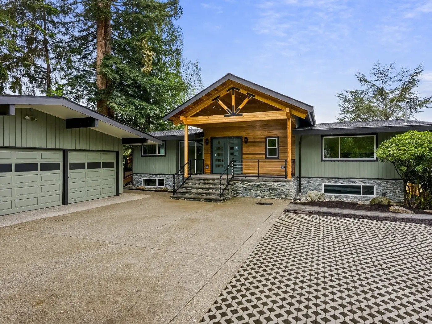 Modern house with a two-car garage, light green siding, wood and stone accents, and a covered entryway with steps; surrounded by trees and a patterned driveway.