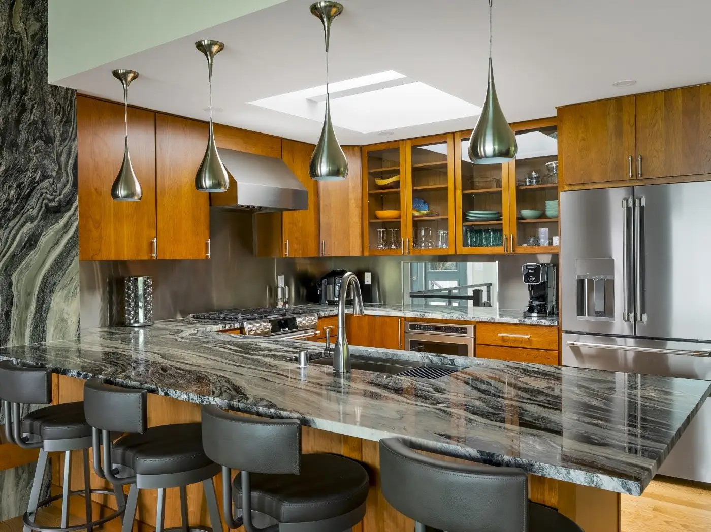 Modern kitchen with wood cabinets, stainless steel appliances, pendant lights, and a large island with a marble countertop and five black barstools. Open shelving displays dishes and glassware.