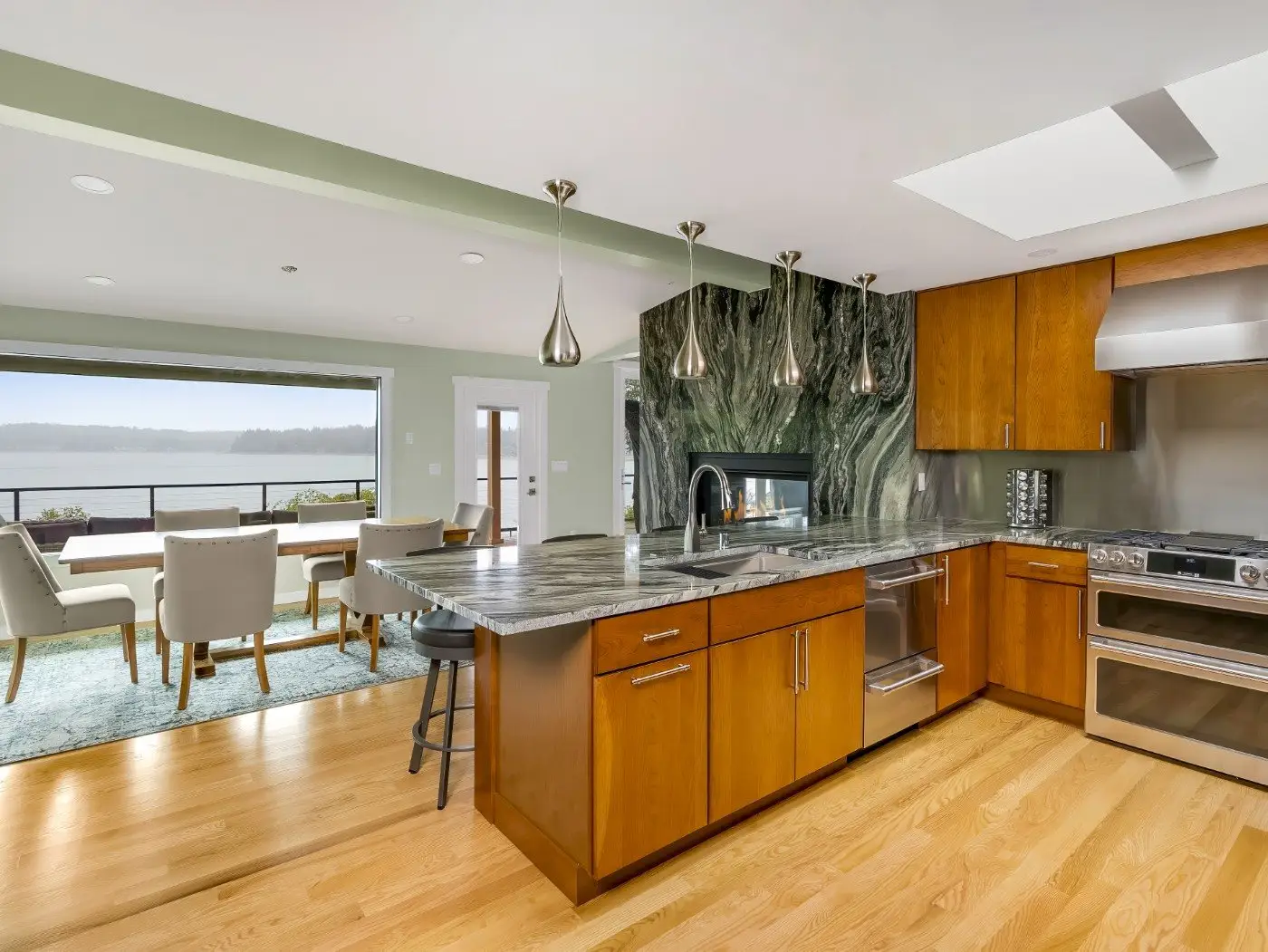 Modern kitchen with wooden cabinets, marble countertops, stainless steel appliances, pendant lights, and an open view of a dining area and large windows overlooking water scenery.