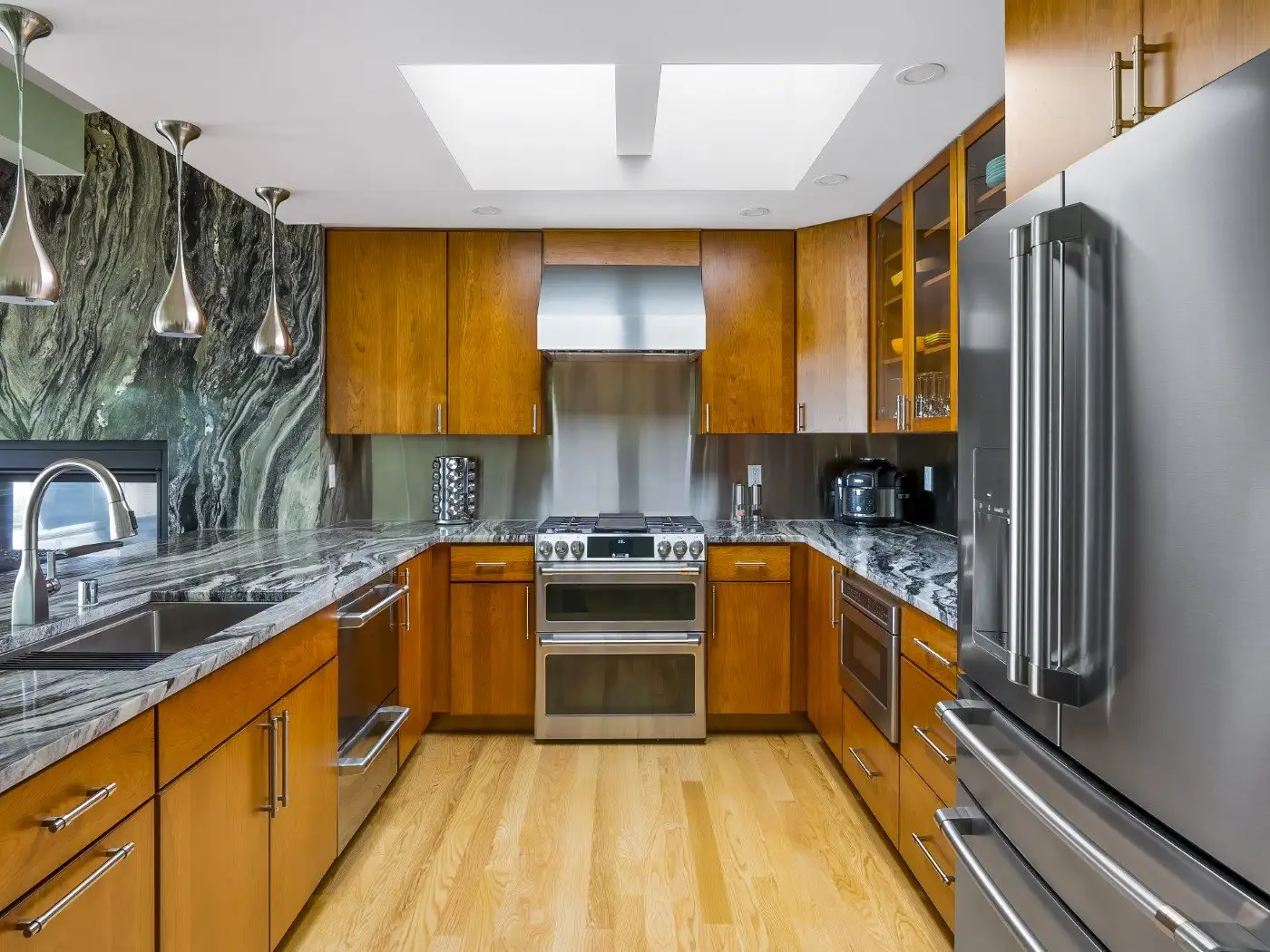 Modern kitchen with wooden cabinets, stainless steel appliances, marble countertops, a double-door refrigerator, and pendant lights. Light wood flooring and a skylight brighten the space.