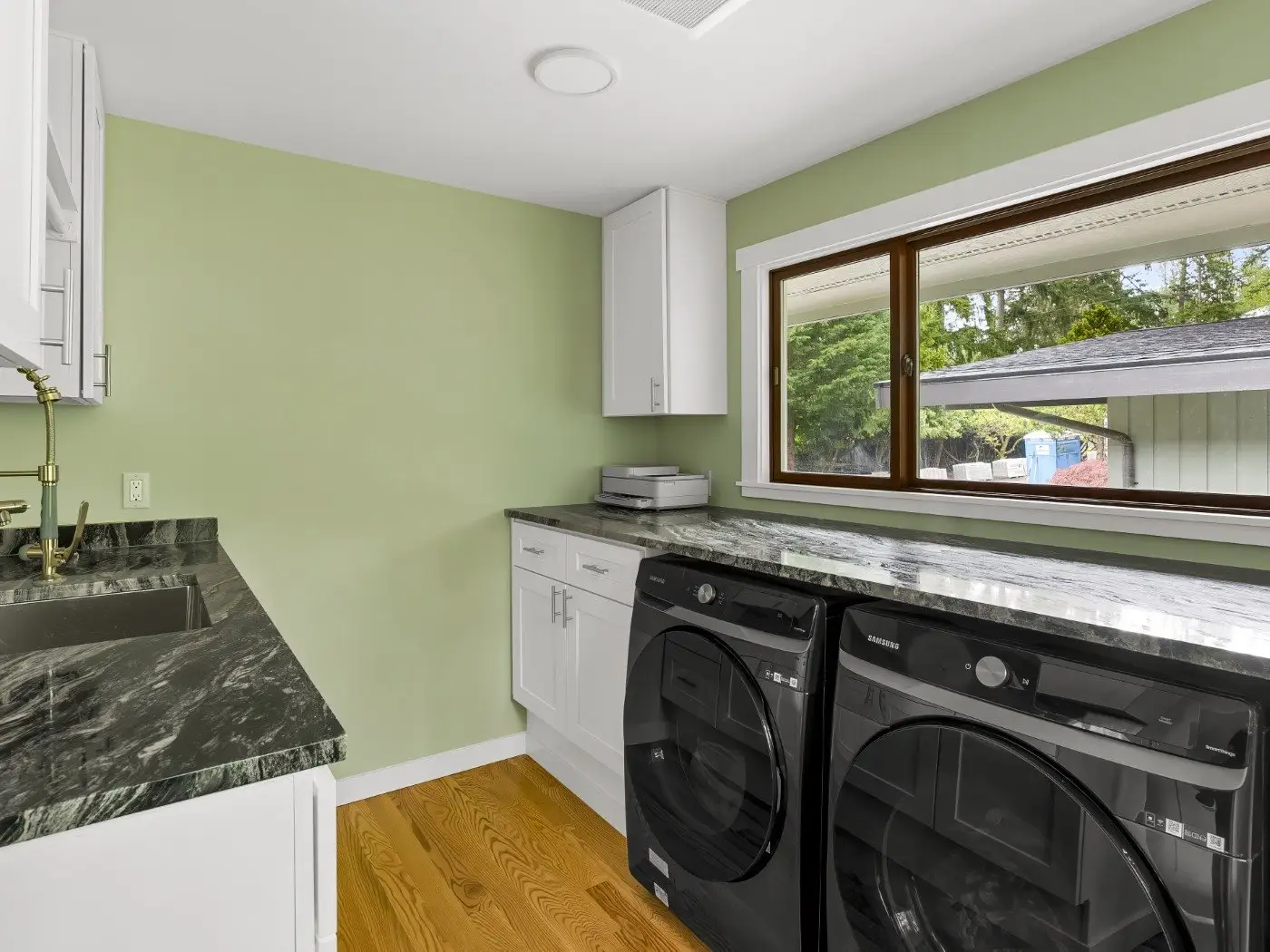 A modern laundry room with light green walls, white cabinets, black countertop, stainless steel sink, and front-loading black washer and dryer under a wide window with a view of trees outside.