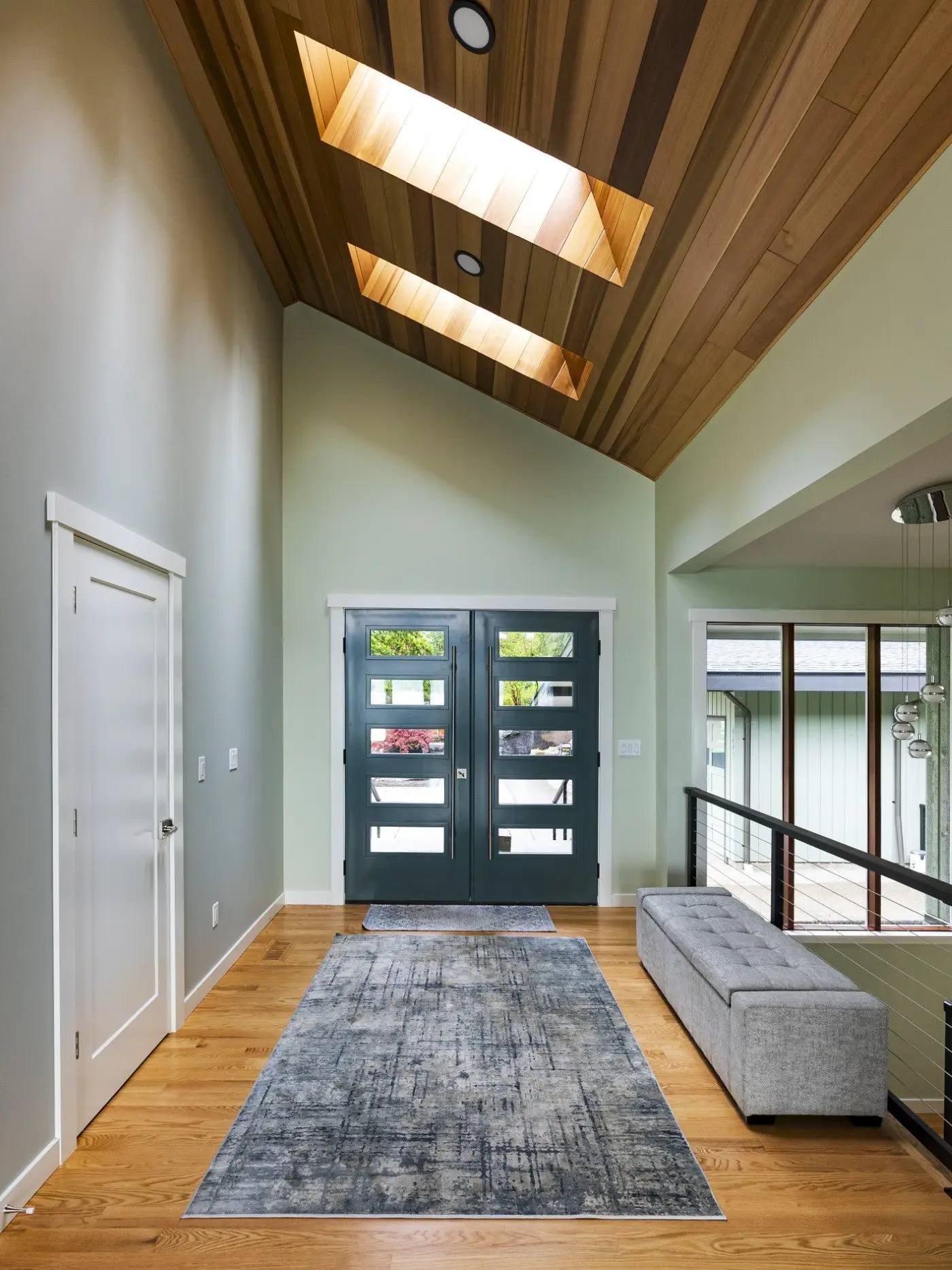 Modern entryway with double teal doors, wooden floor, and a blue-gray rug. The ceiling is angled and paneled with wood, featuring two skylights. A gray bench sits to the right, near metal railing and large windows.