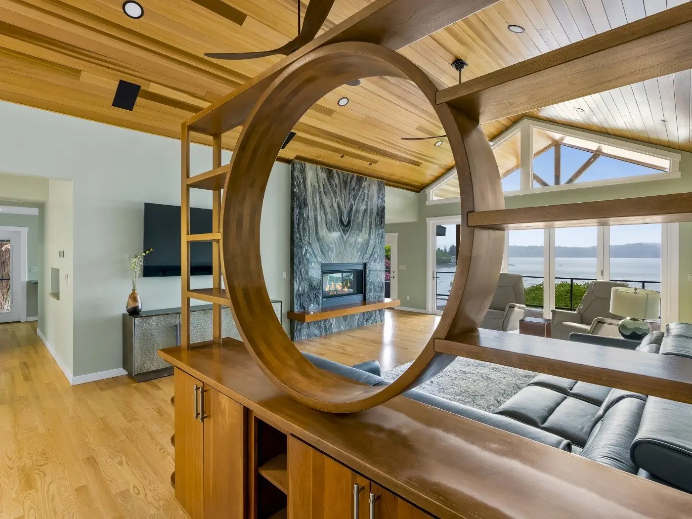 Modern living room with wood floors and ceiling, a large circular wooden divider, blue sectional sofa, fireplace with marble surround, and large windows offering a scenic view of water and trees.