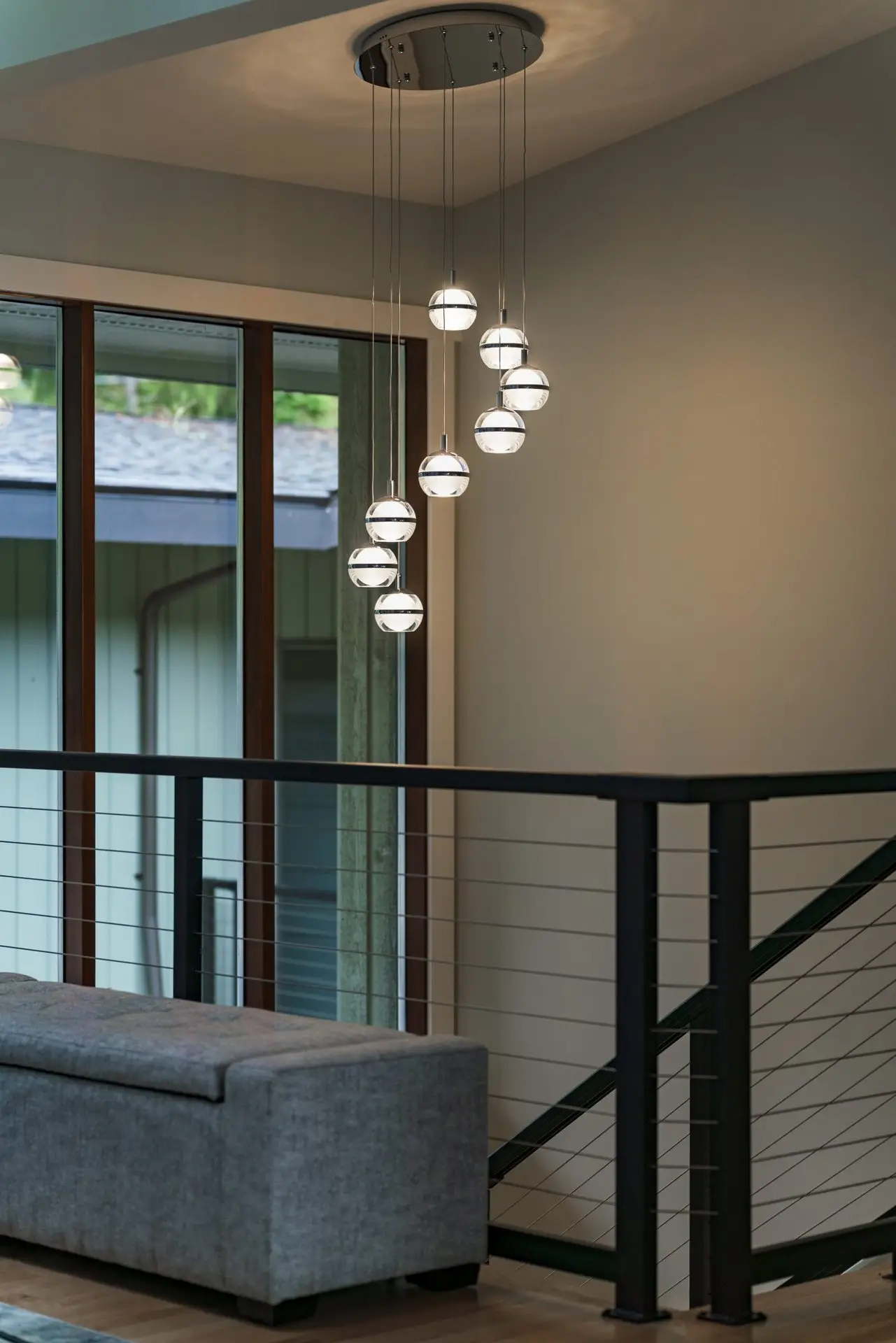 A modern, multi-globe pendant light hangs above a staircase with metal railings in a contemporary home, next to large windows and a gray sofa.