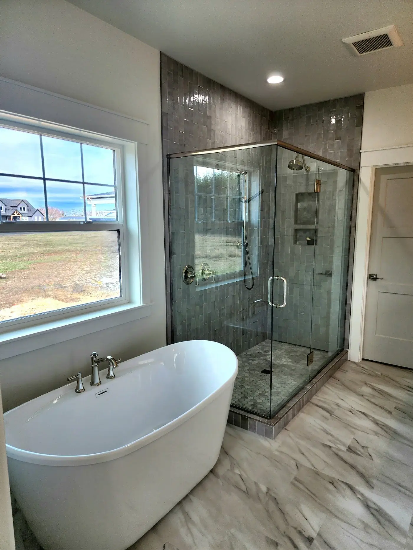 A modern bathroom with a freestanding white bathtub next to a large window, and a glass-enclosed shower with gray tiles and a rainfall showerhead. The floor has white and gray marble-patterned tiles.