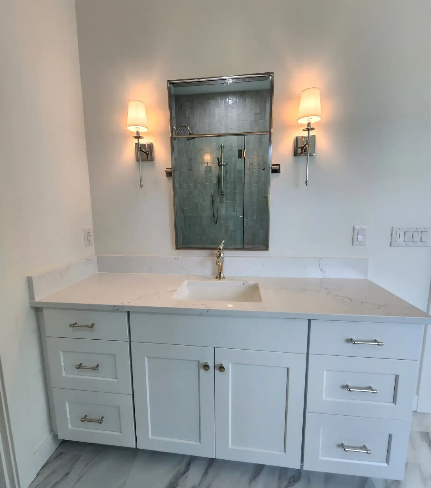 A modern bathroom vanity with a white countertop and sink, white cabinets, a wall-mounted mirror reflecting a shower, and two wall sconces with white shades on either side of the mirror.