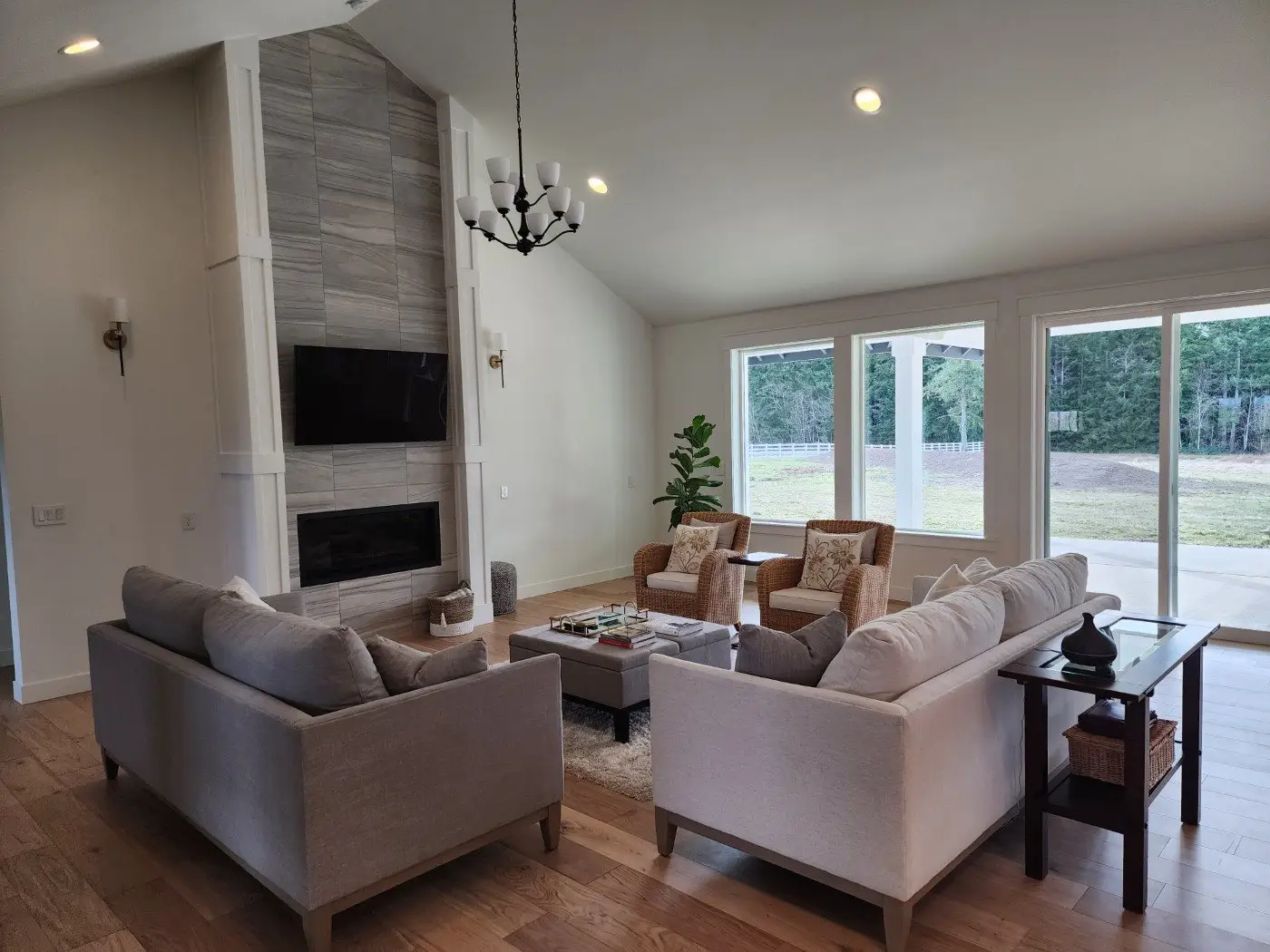 A modern living room with light-colored sofas, armchairs, a coffee table, a fireplace with a mounted TV, and large windows showing a backyard and trees outside. The room has wooden floors and neutral decor.