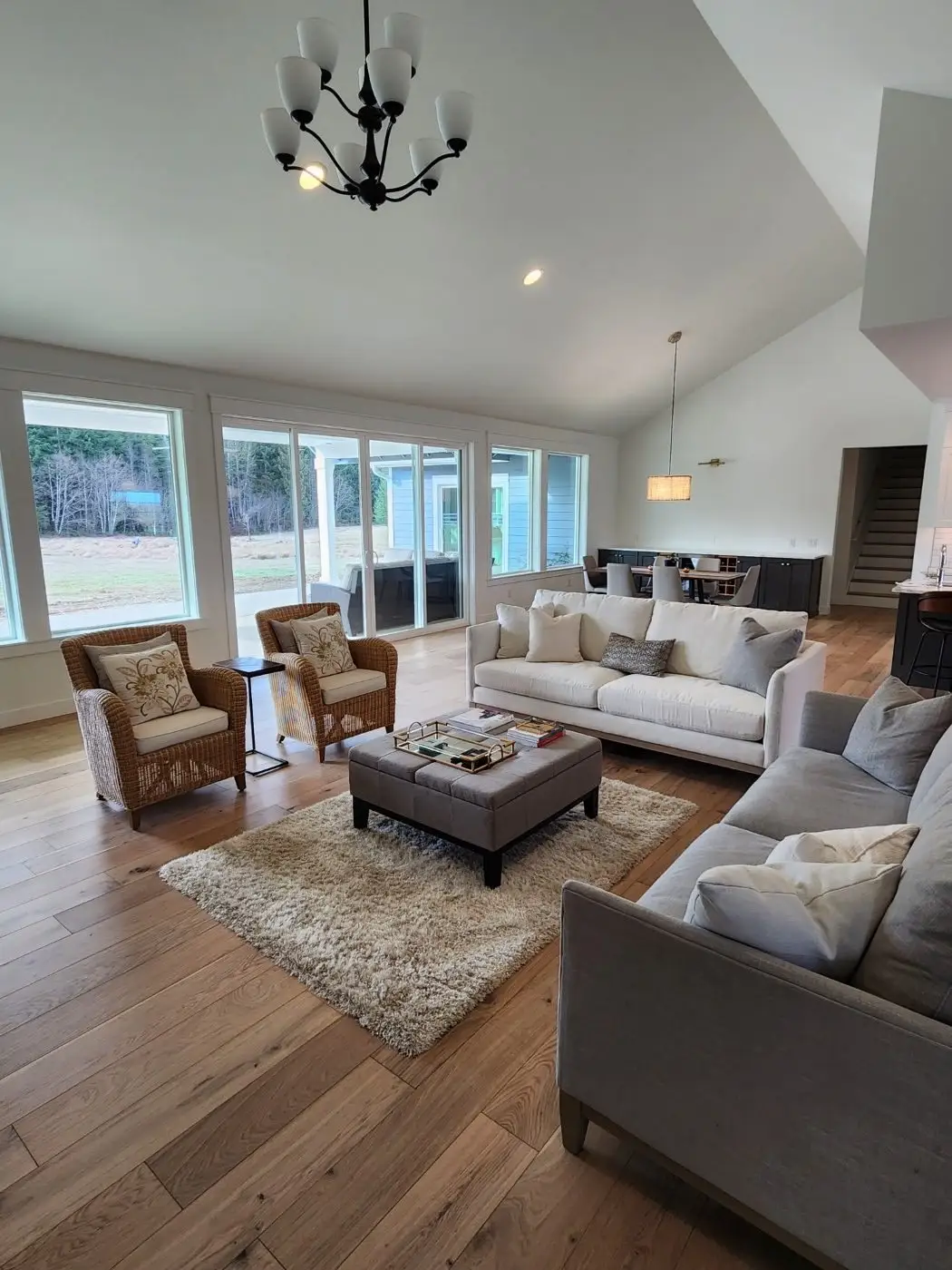 Spacious living room with large windows, two sofas, wicker chairs, a plush rug, coffee table, and light wood floors. Chandelier and pendant lights hang from high ceilings; outdoor view visible through windows.