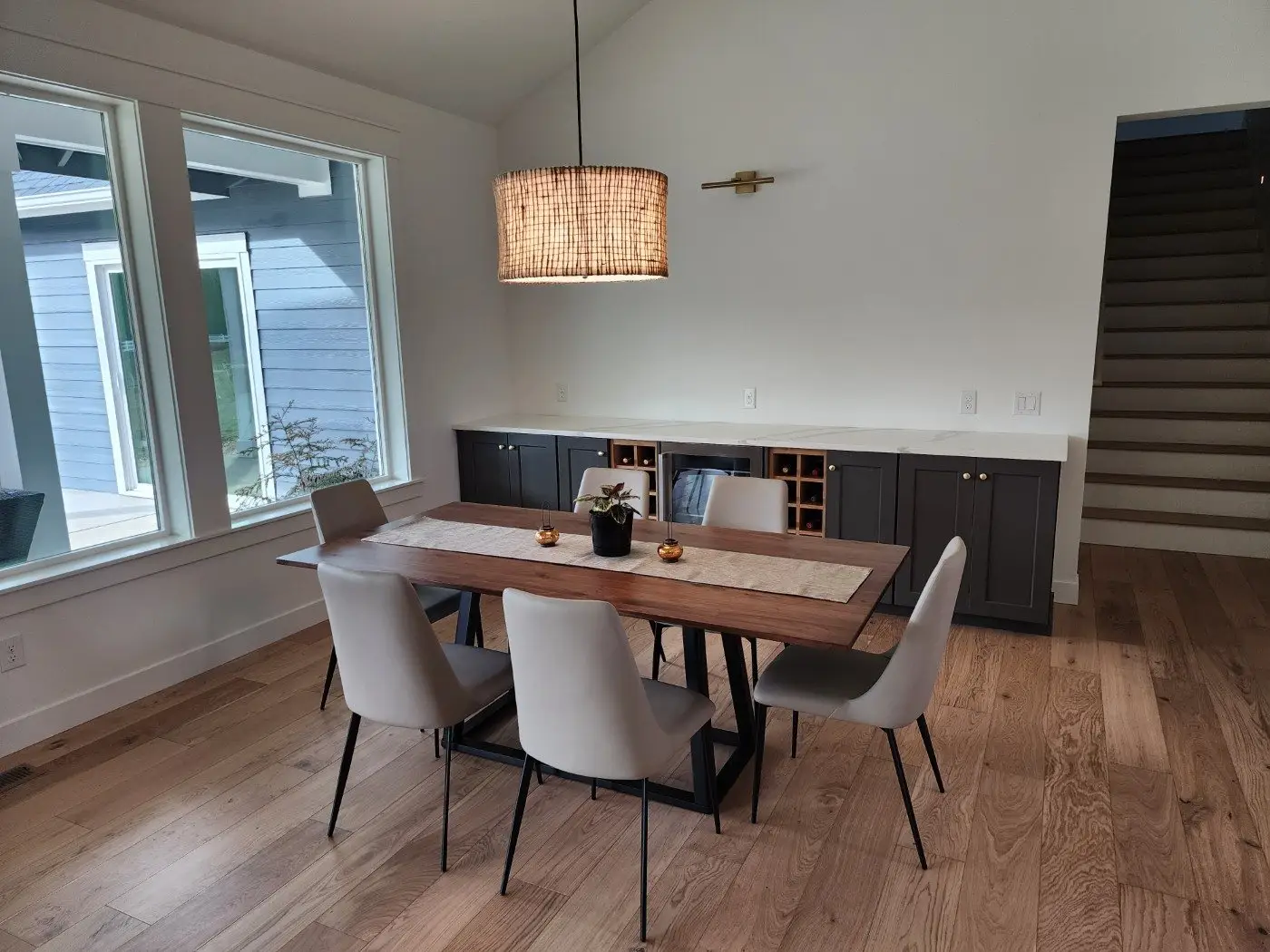 A modern dining room with a wooden table, six beige chairs, a woven pendant light, built-in cabinets, and a large window letting in natural light. A staircase is visible nearby.