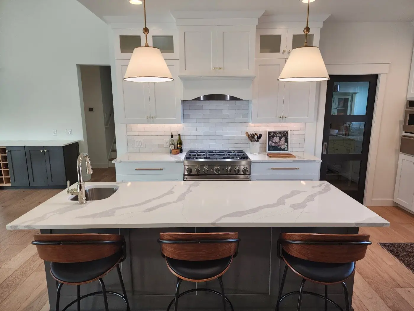 Modern kitchen with a large white marble island, three black and wood barstools, pendant lights, stainless steel appliances, white cabinets, and a subway tile backsplash. A sink and faucet are on the island.