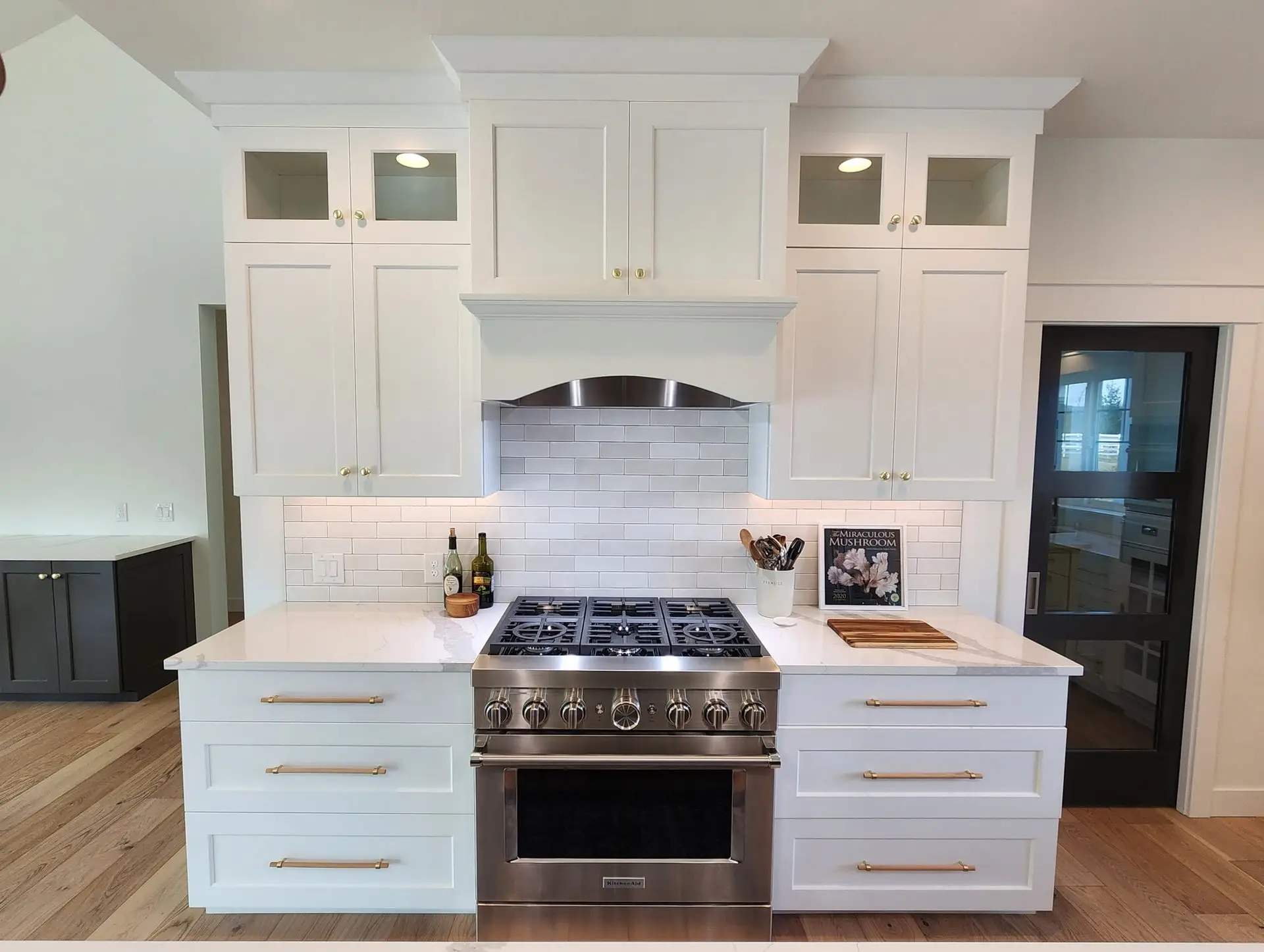 Modern white kitchen with a stainless steel gas stove, white cabinets, subway tile backsplash, and marble countertop. Bottles and a cutting board sit on the counter next to a cookbook stand.