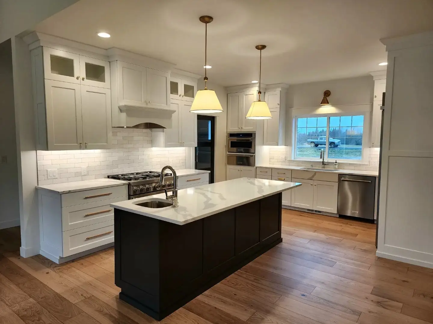 Modern kitchen with white cabinets, stainless steel appliances, a large dark island with a sink, pendant lights above, white tiled backsplash, and wooden floors. A window above the sink shows an outdoor scene.