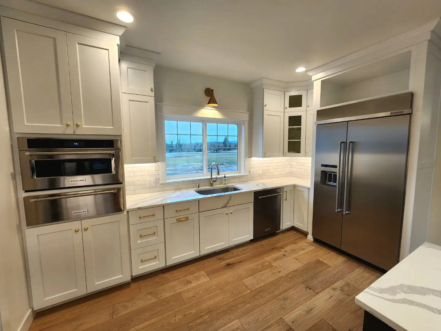 Modern kitchen with white cabinets, stainless steel appliances, a double oven, large refrigerator, dishwasher, farmhouse sink under a window, and wooden floors. White subway tile backsplash and under-cabinet lighting are visible.