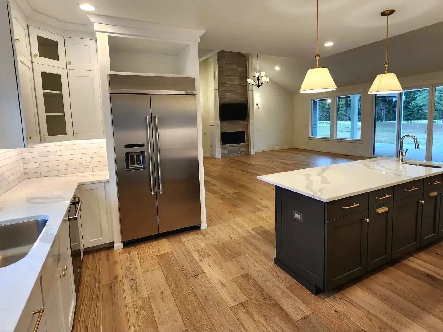 Modern kitchen with stainless steel refrigerator, white cabinets, subway tile backsplash, center island with dark base and white countertop, pendant lights, and open view to a spacious living area with fireplace and large windows.