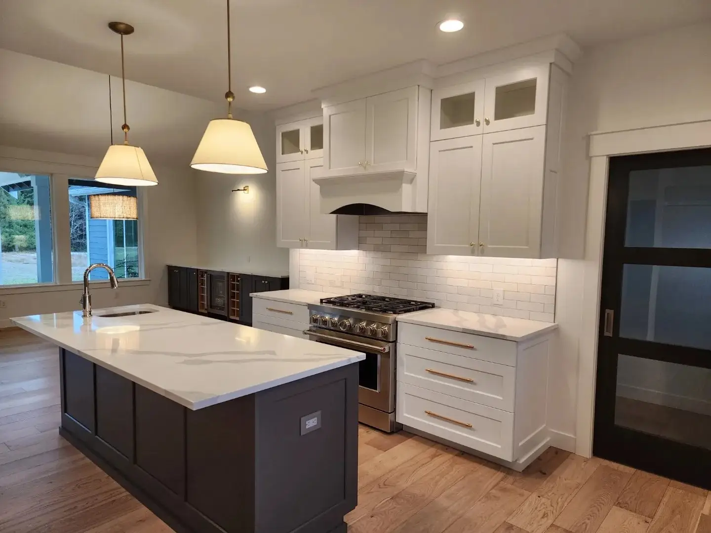Modern kitchen with white cabinets, gold handles, a large island with a sink, pendant lights, a stainless steel stove, subway tile backsplash, wood flooring, and a sliding glass door.