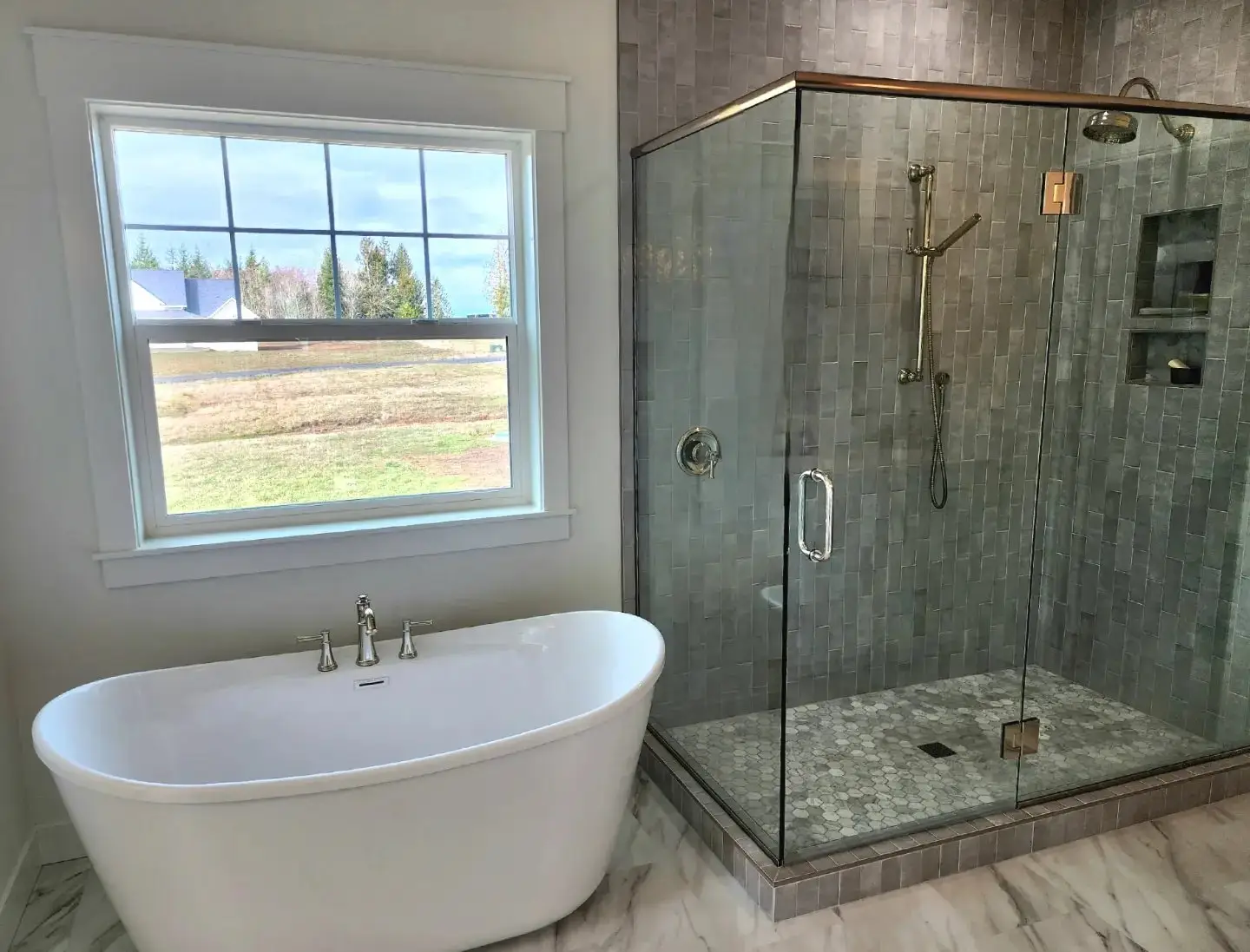 Modern bathroom with a freestanding white bathtub near a large window and a glass-enclosed shower with gray tiles and chrome fixtures on the right.