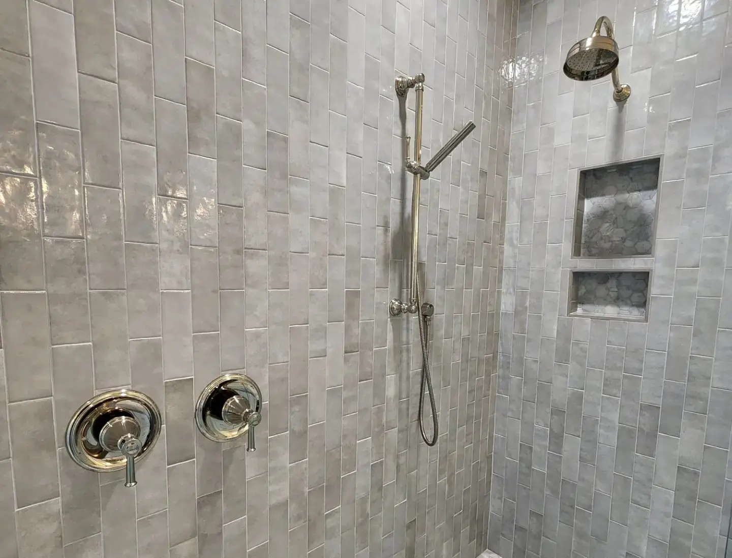 A modern shower with light gray rectangular tiles, two chrome control knobs, a handheld showerhead, a fixed showerhead above, and two recessed shelves in the wall.