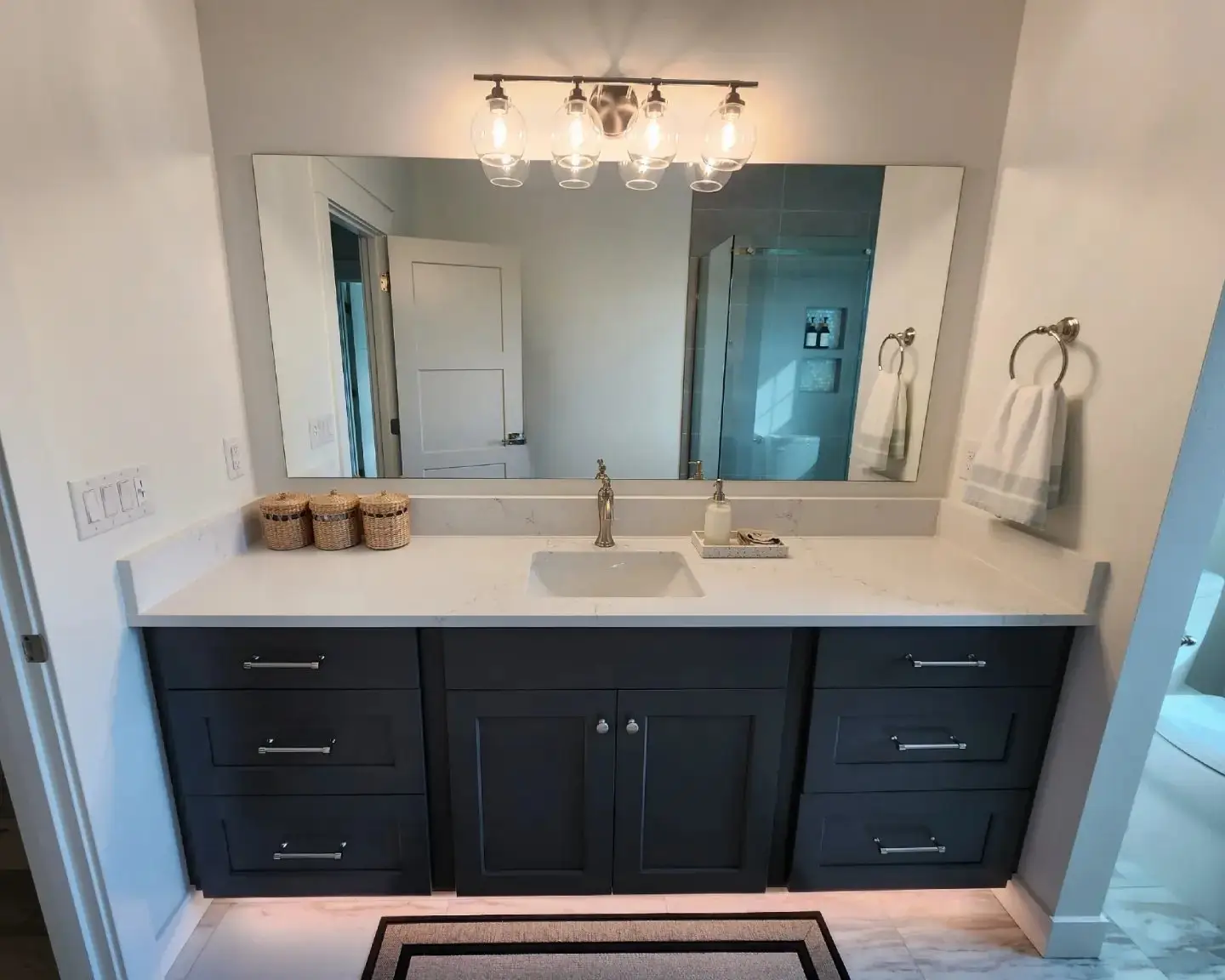 A modern bathroom vanity with dark cabinets, a white countertop, a built-in sink, a large mirror, and a three-light fixture above. Three small baskets, a towel ring, and a tray with soap are on the counter.