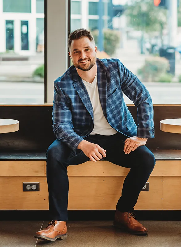 Daniel Doyle, founder of Doyle Construction, seated on a wooden bench in a blue plaid blazer