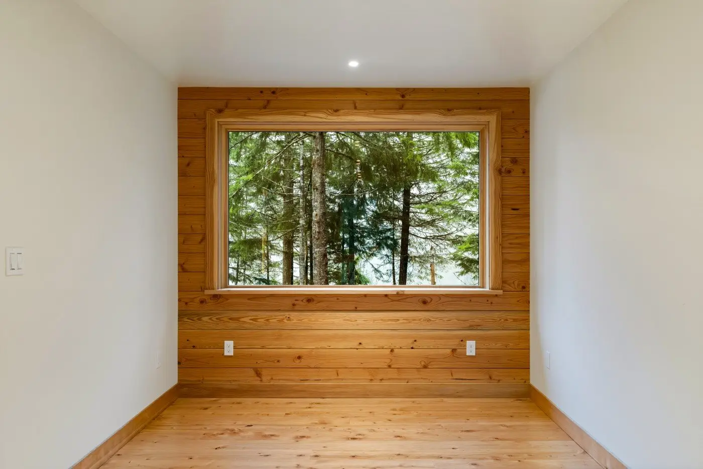 A small empty room with light wood floors and walls, featuring a large window framed in wood. The window overlooks tall evergreen trees outside, letting in natural light.