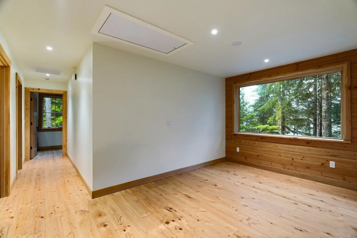 A bright, empty room with light wood floors and walls, large window with a forest view, recessed lighting, and a hallway leading to another area.