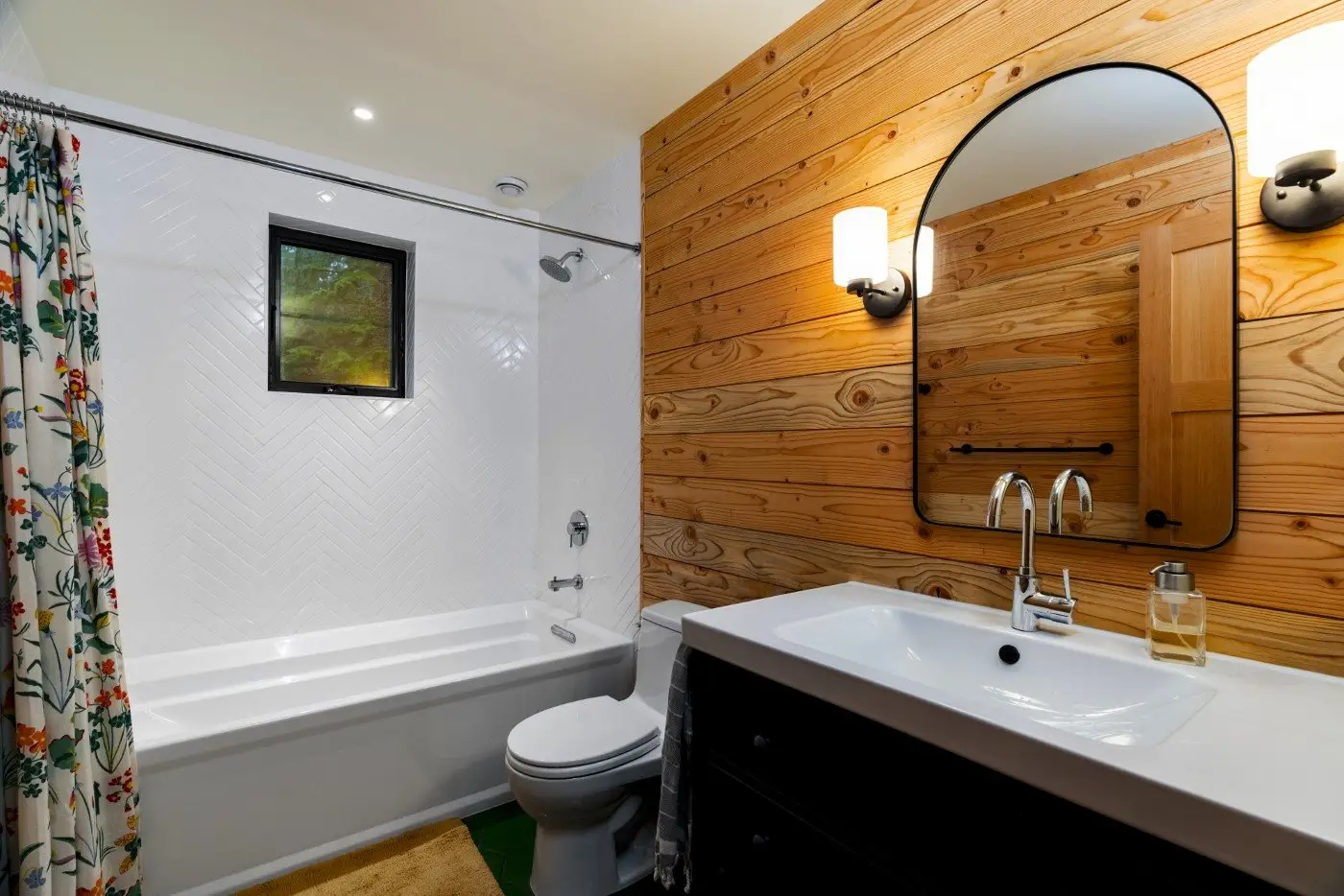 A bathroom with wood-paneled walls, a white bathtub with a floral shower curtain, a small window, a white sink with a black vanity, a modern oval mirror, and two wall-mounted light fixtures.