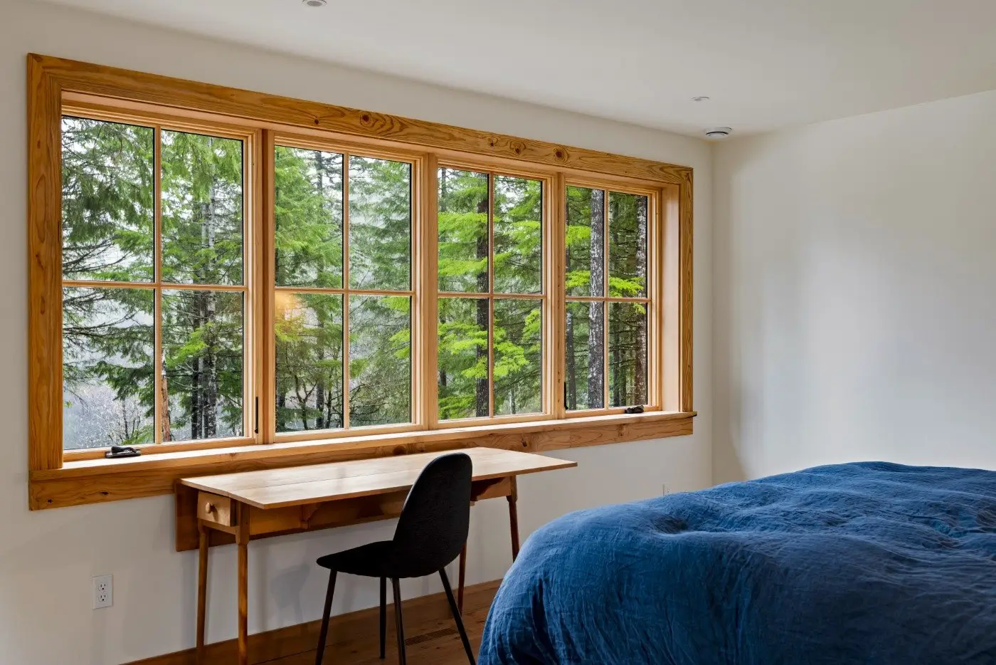 A simple bedroom with a wooden desk and black chair by a large window overlooking green trees, next to a bed covered with a blue comforter. Natural light fills the room, creating a calm and cozy atmosphere.