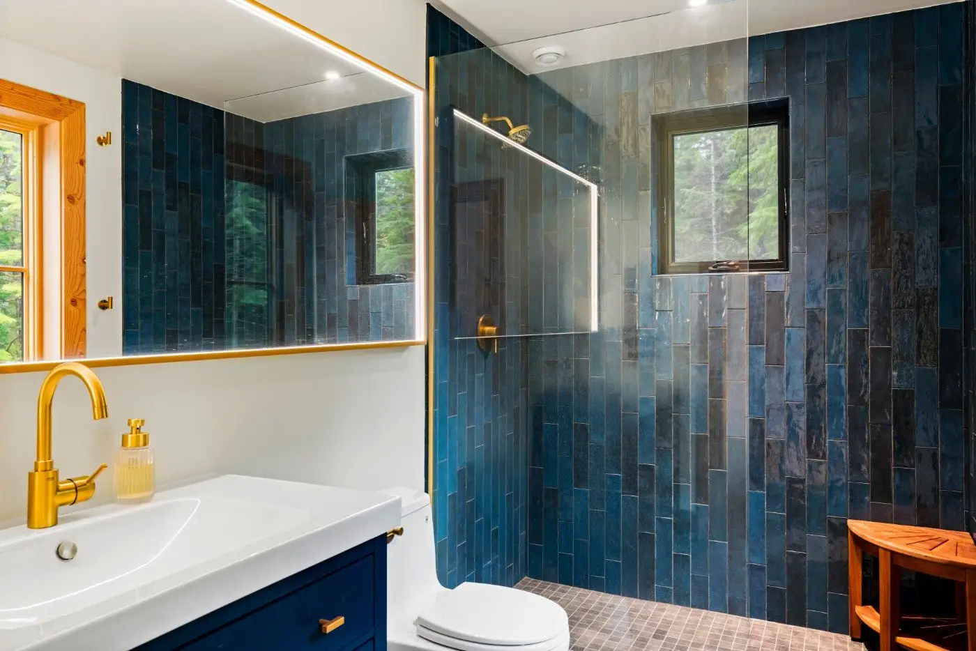 Modern bathroom with deep blue tile shower, gold fixtures, white sink, wood-framed mirror, small window, and a wooden stool. The shower has a glass partition and natural light comes in through the window.