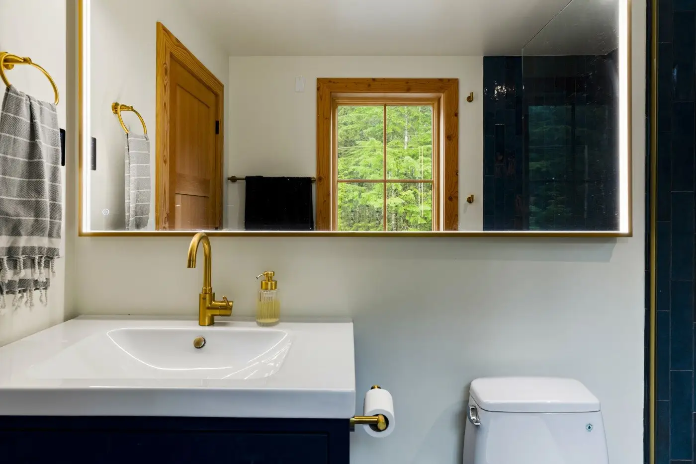 A bathroom with a white sink, gold faucet, striped towel, soap dispenser, and toilet. A large mirror reflects a wooden-framed window showing green trees outside. The door and towel rack are wooden.