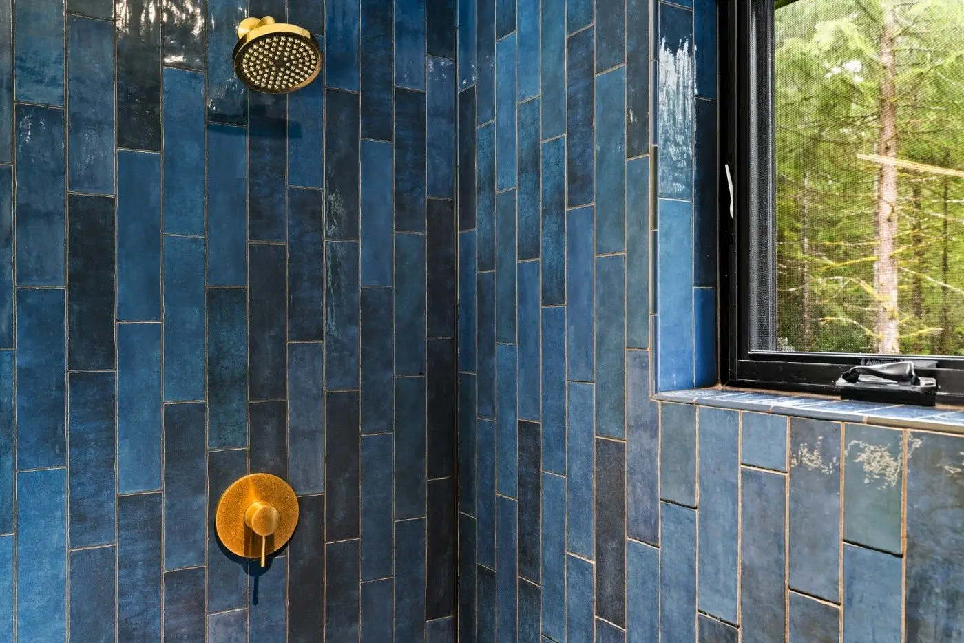 Blue-tiled shower with gold showerhead and control handle, next to a window showing green trees outside. Natural light illuminates the space.