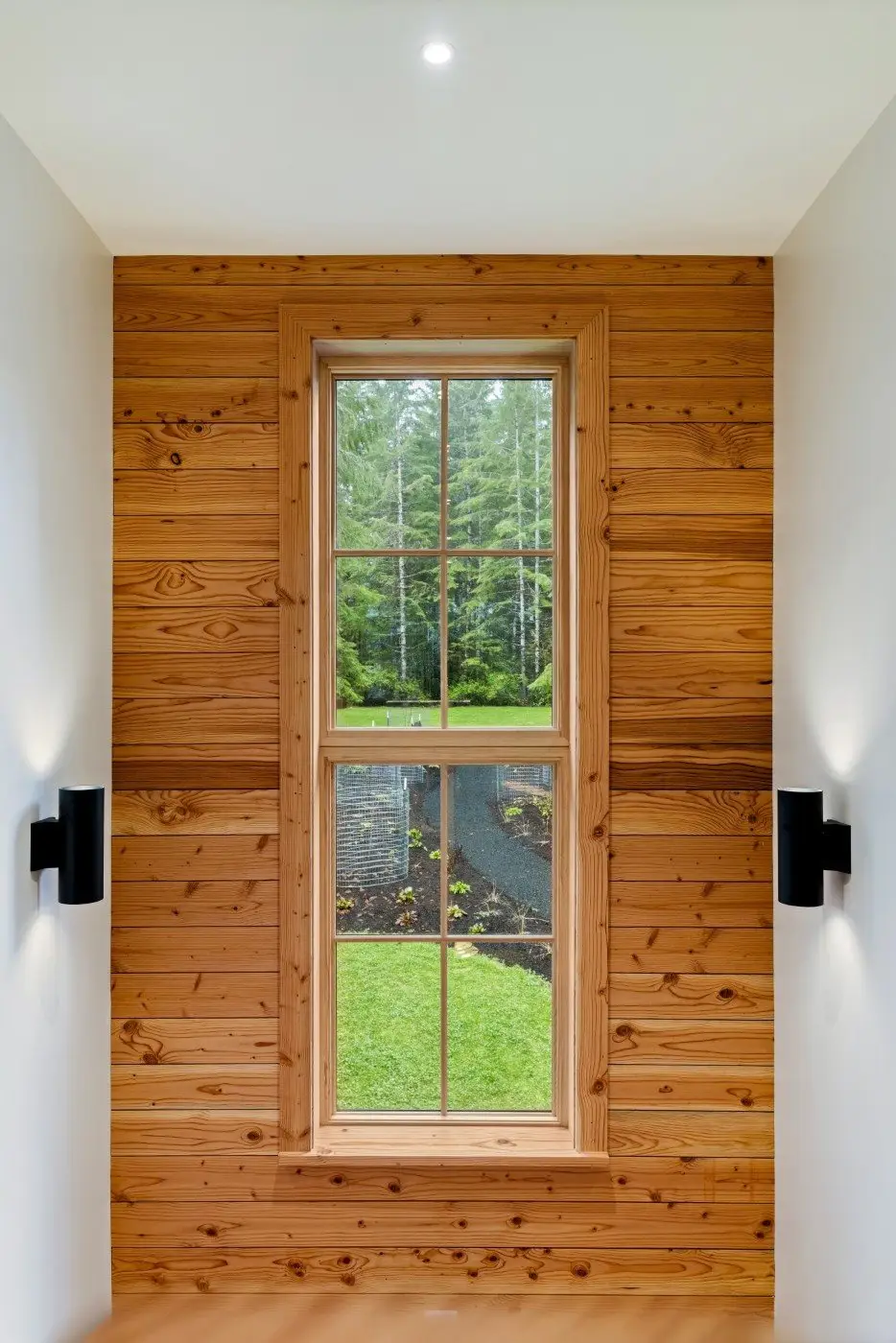 A tall wooden-framed window set in a wall of horizontal wood panels, looking out onto a green lawn and trees. Two modern black wall lights are mounted on either side of the window.