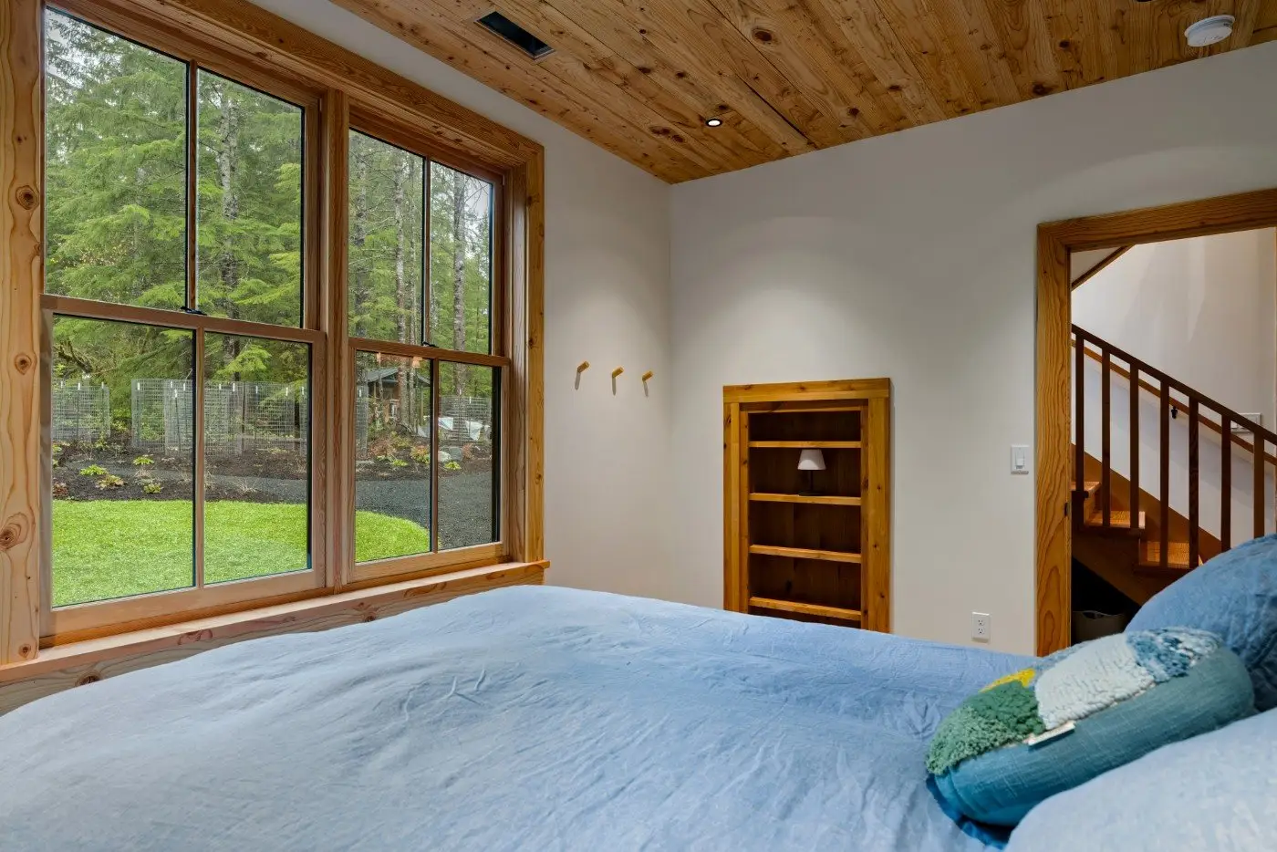 A cozy bedroom with a large window showing a green forested yard, light wood ceiling and trim, a blue bedspread, a small built-in bookshelf, and a staircase seen through an open doorway.