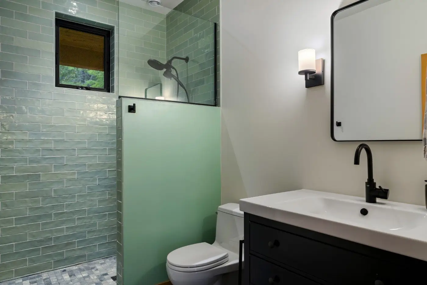 Modern bathroom with green tiled walk-in shower, black showerhead, partial glass divider, white toilet, white sink with black faucet, black vanity, and wall-mounted light next to a rectangular mirror.