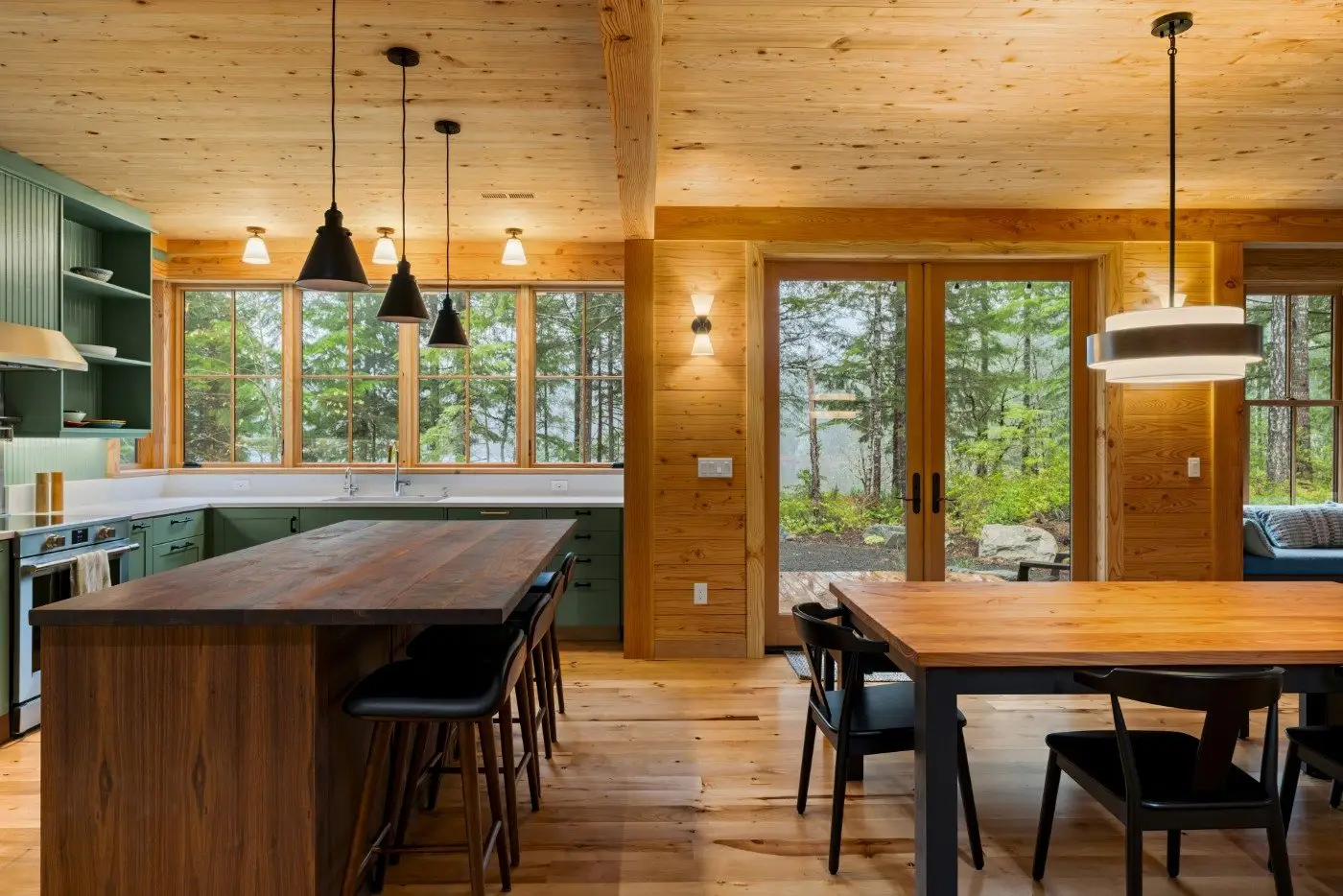 Modern rustic kitchen and dining area with wood-paneled walls, large windows, green cabinets, a wooden island with bar stools, hanging pendant lights, and glass doors overlooking a forested yard.