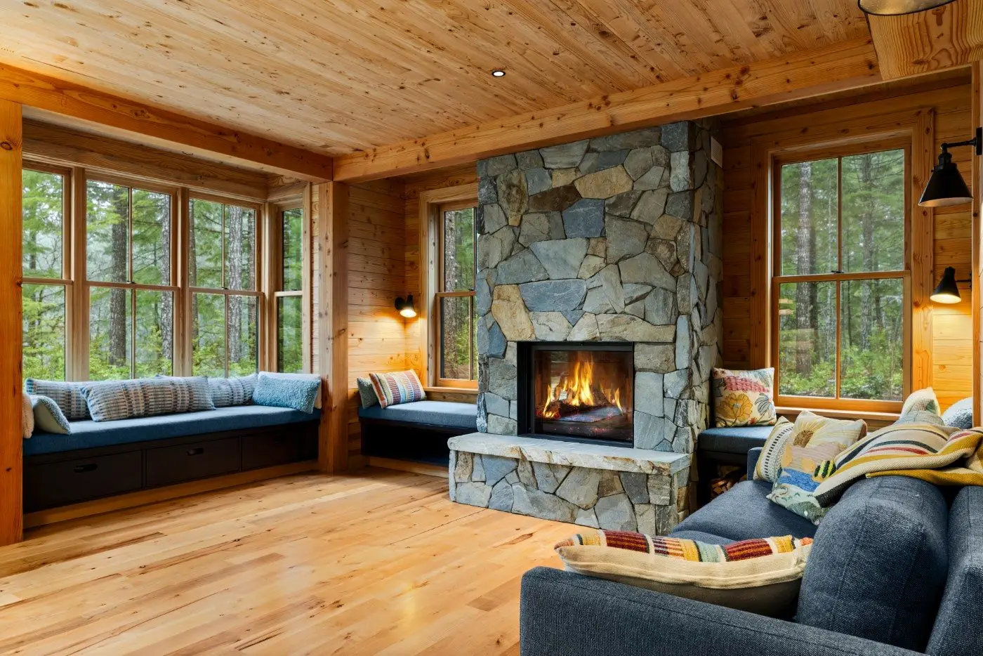 A cozy living room with wood-paneled walls and floor, large windows with forest views, a stone fireplace with a burning fire, built-in window seats, and a blue sofa with colorful pillows.