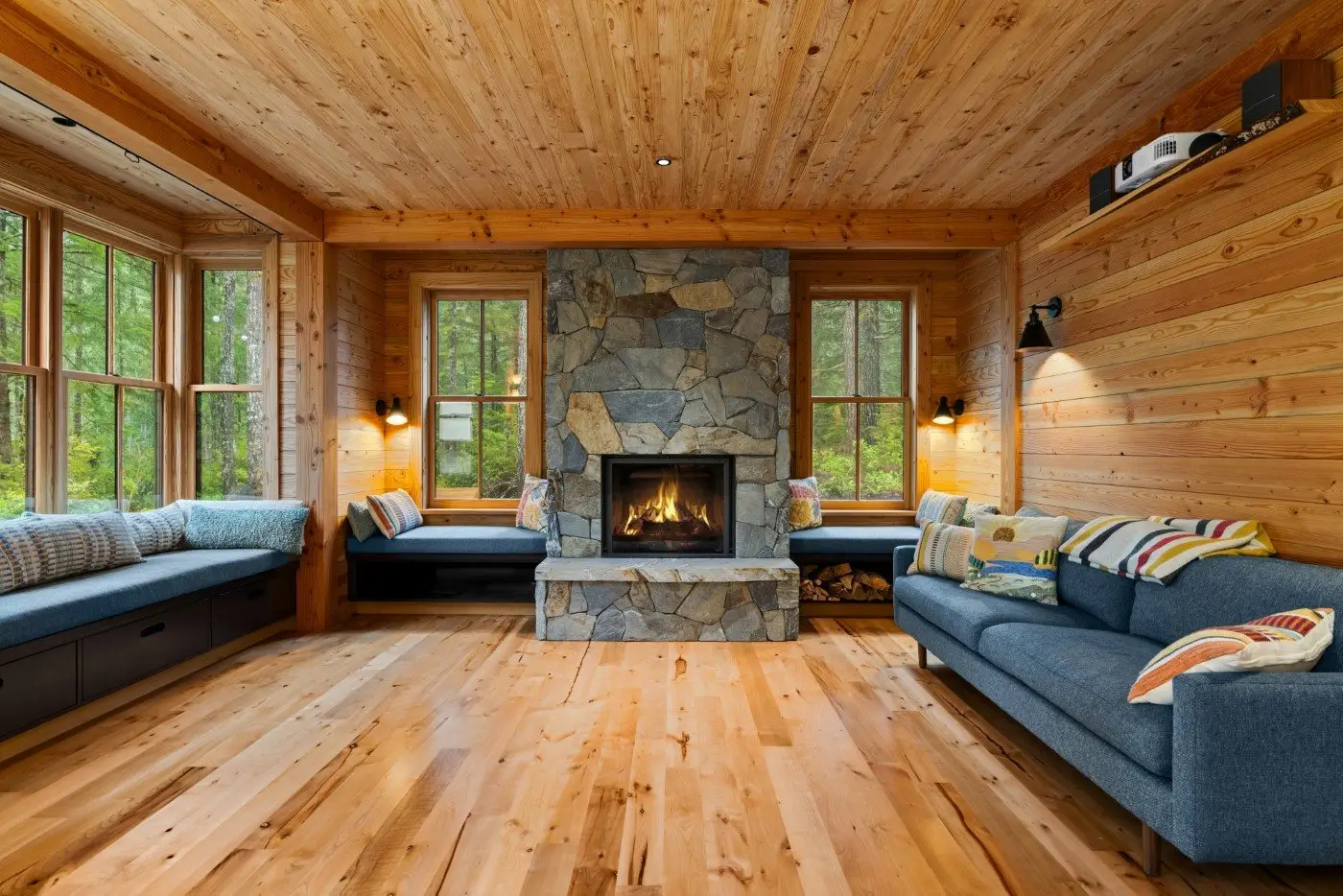Cozy living room with wood-paneled walls and ceiling, a stone fireplace in the center, blue sofas, window seats with cushions, and large windows showing green forest outside.