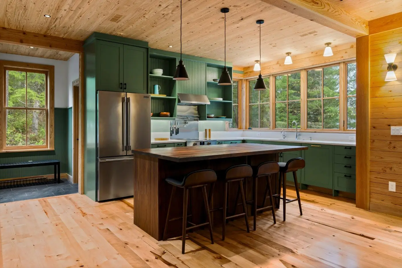 A modern kitchen with green cabinets, a stainless steel fridge, a dark wood island with four bar stools, pendant lights, large windows, and light wood floors and ceiling.