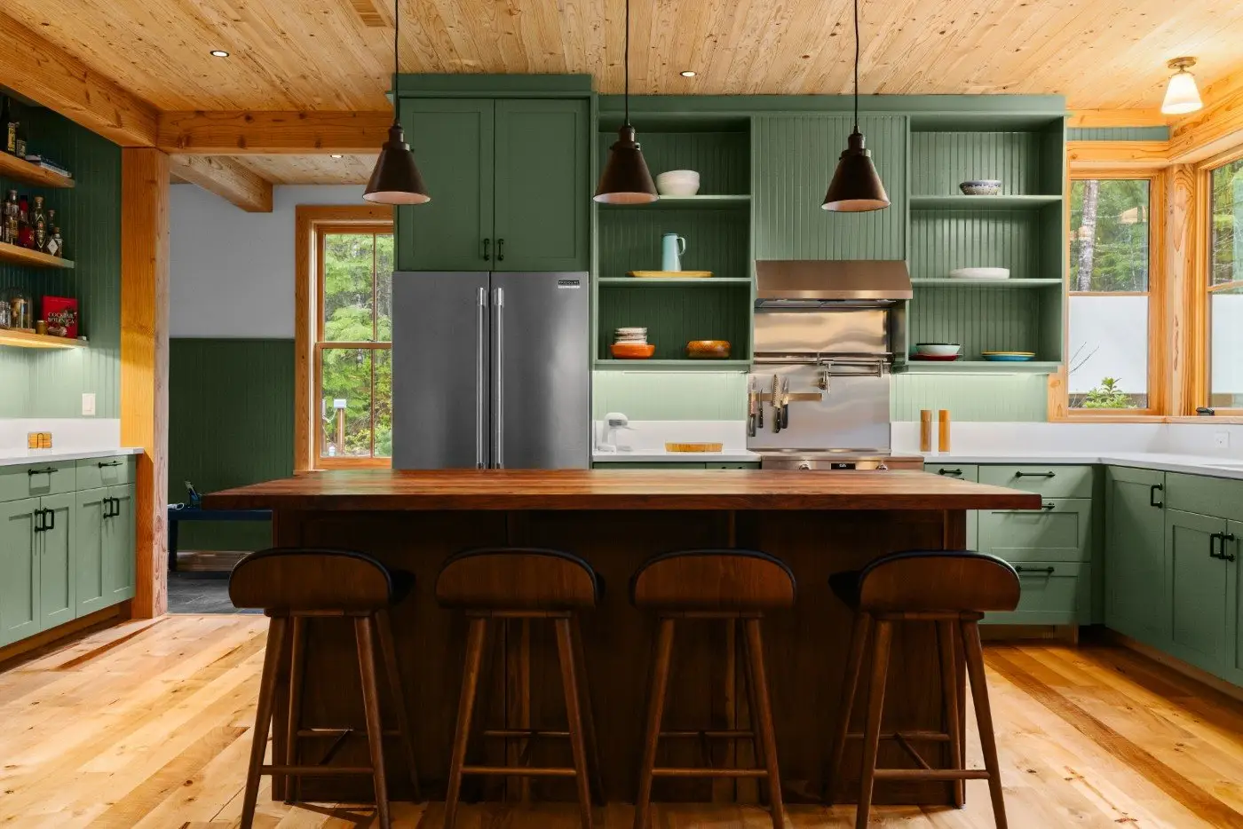 A modern kitchen with sage green cabinets, wood accents, a wooden island with four bar stools, stainless steel appliances, pendant lights, and large windows letting in natural light.