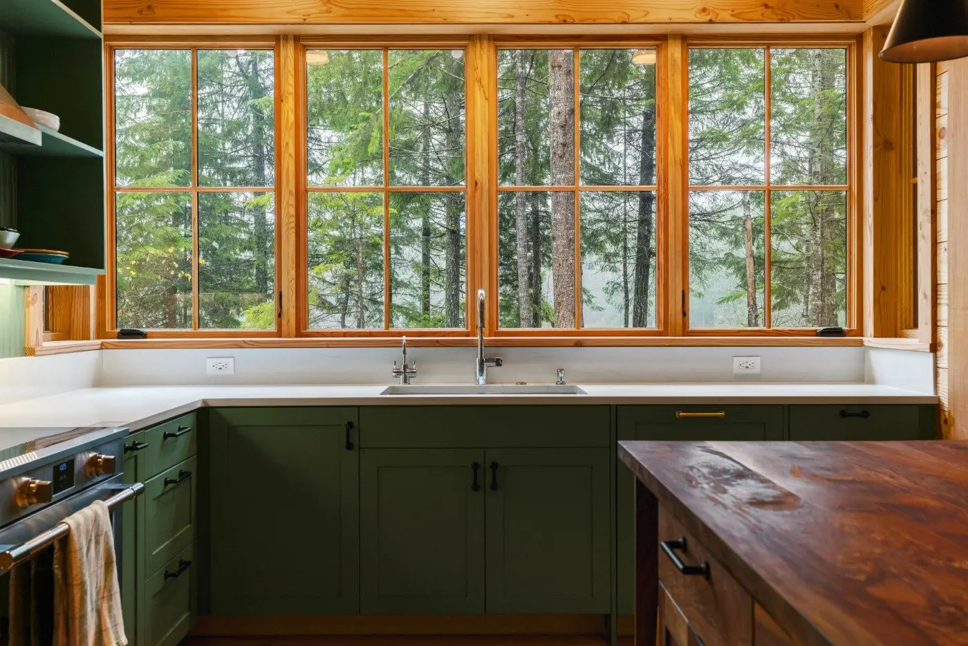 A bright kitchen with green cabinets, a large farmhouse sink, wooden countertops, and wide windows offering a view of tall trees and greenery outside.