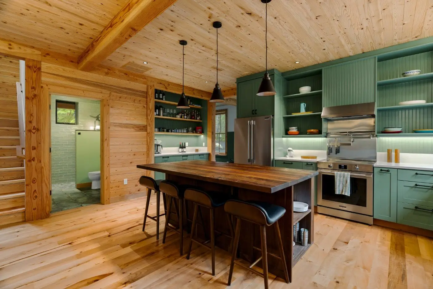 A modern kitchen with wood paneling, green cabinets, stainless steel appliances, and a wooden island with four black barstools; open shelves display dishes, and a doorway leads to a bathroom.