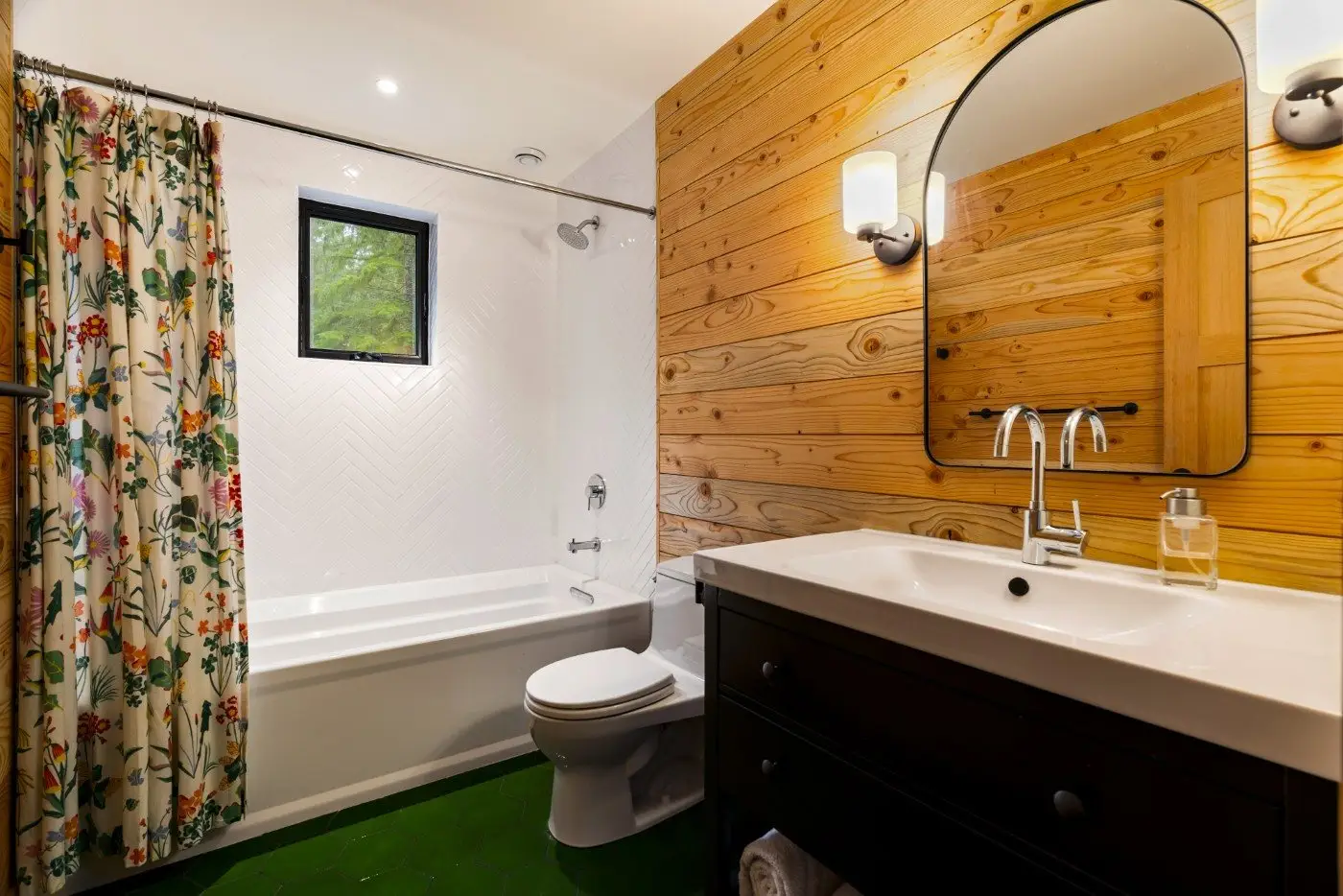 A bathroom with a floral shower curtain, white bathtub, toilet, dark wood vanity with a white sink, oval mirror, wall sconce, and wood-paneled wall. There is a small window above the tub and green floor tiles.