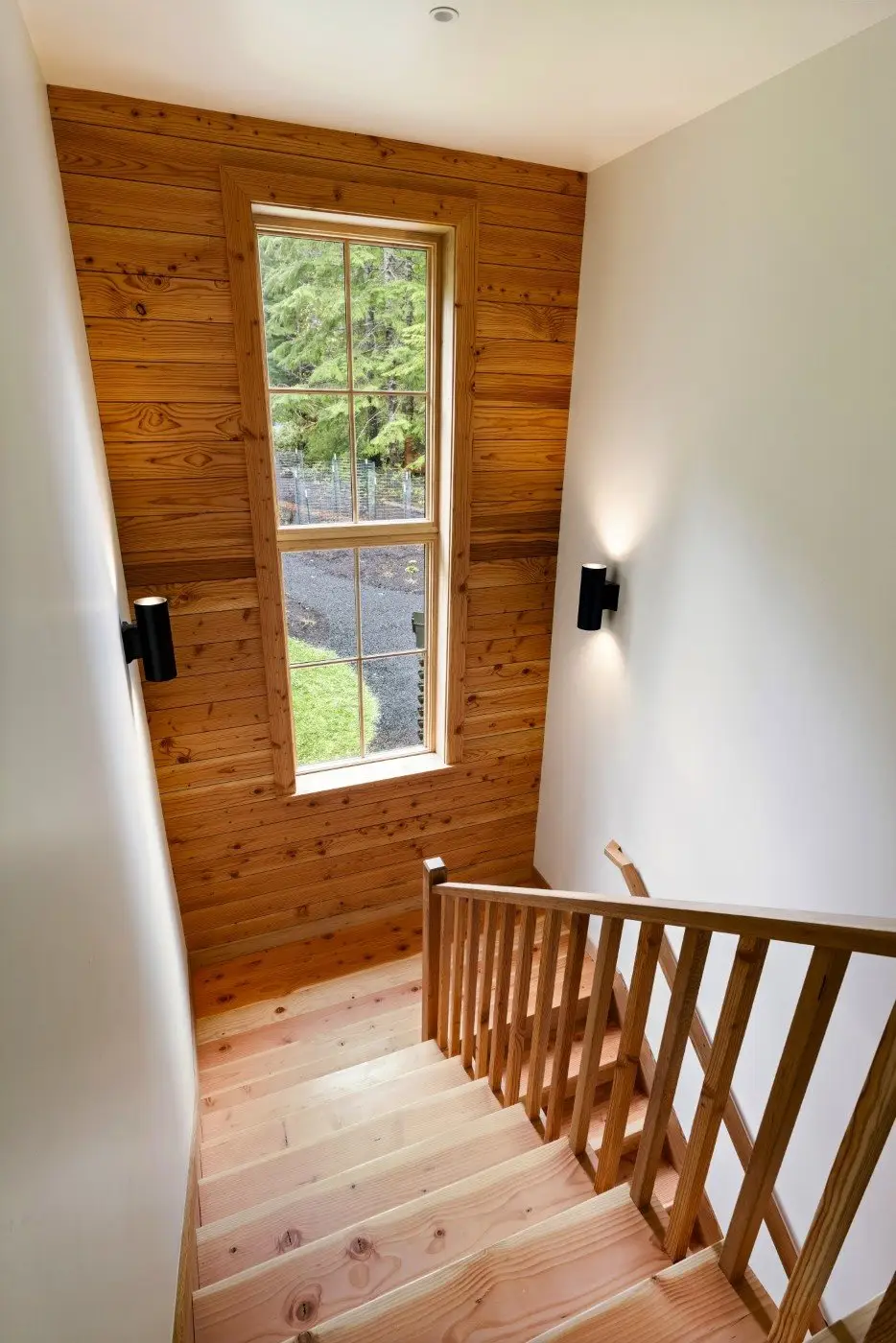 A wooden staircase with matching railing leads down to a landing beneath a tall window. Natural light fills the space, highlighting the wood-paneled wall and light fixtures on either side. Trees and grass are visible outside.