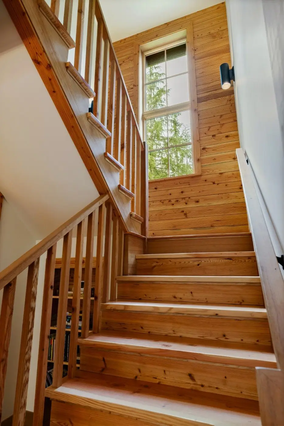 A wooden staircase with railings leads up to a landing with a large window. Sunlight shines through the window, illuminating the warm wood tones and a view of green trees outside.