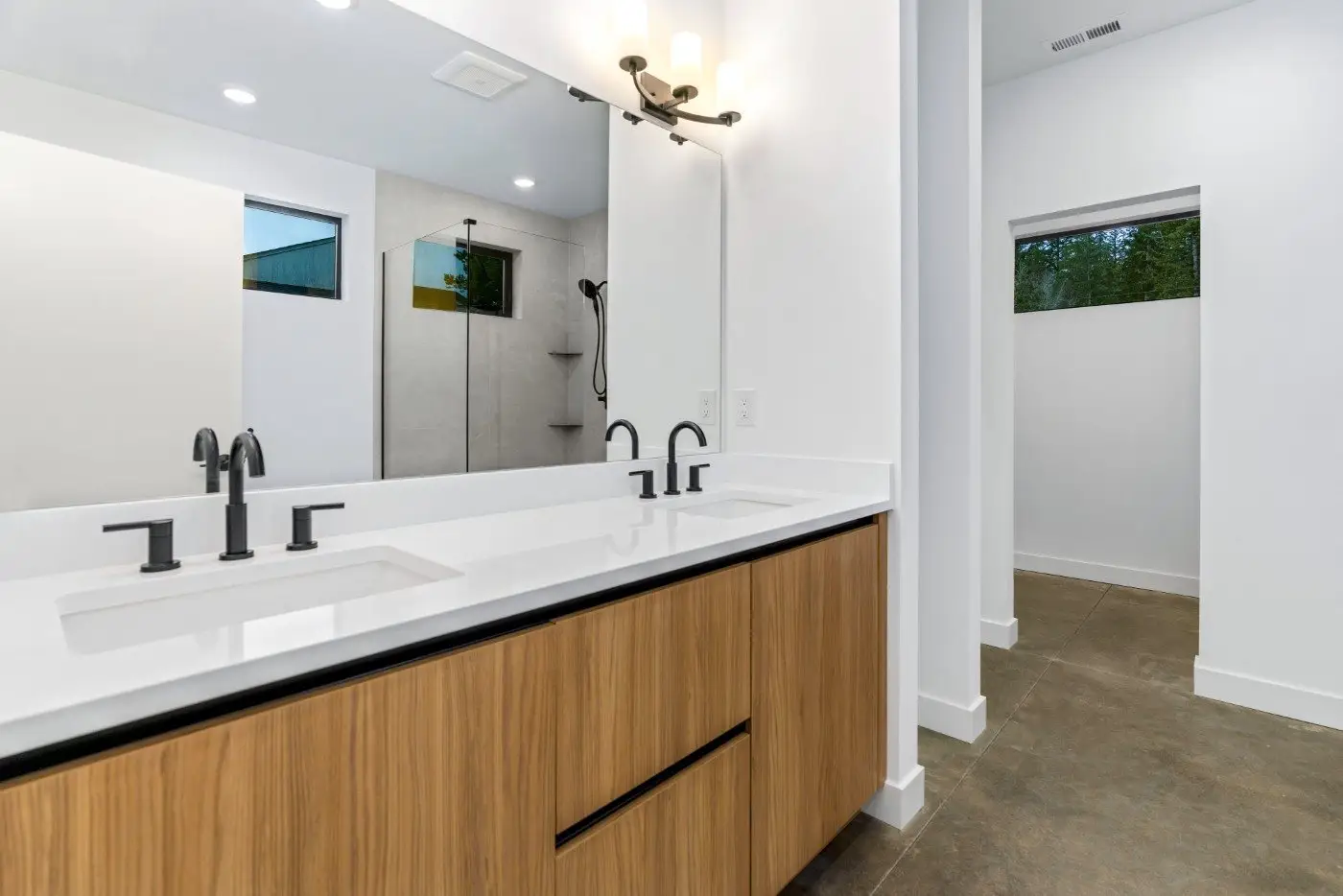 Modern bathroom with double sink vanity, black faucets, large mirror, light fixture above, and a walk-in shower with glass panel in the background. Neutral tones and clean, minimalist design throughout.