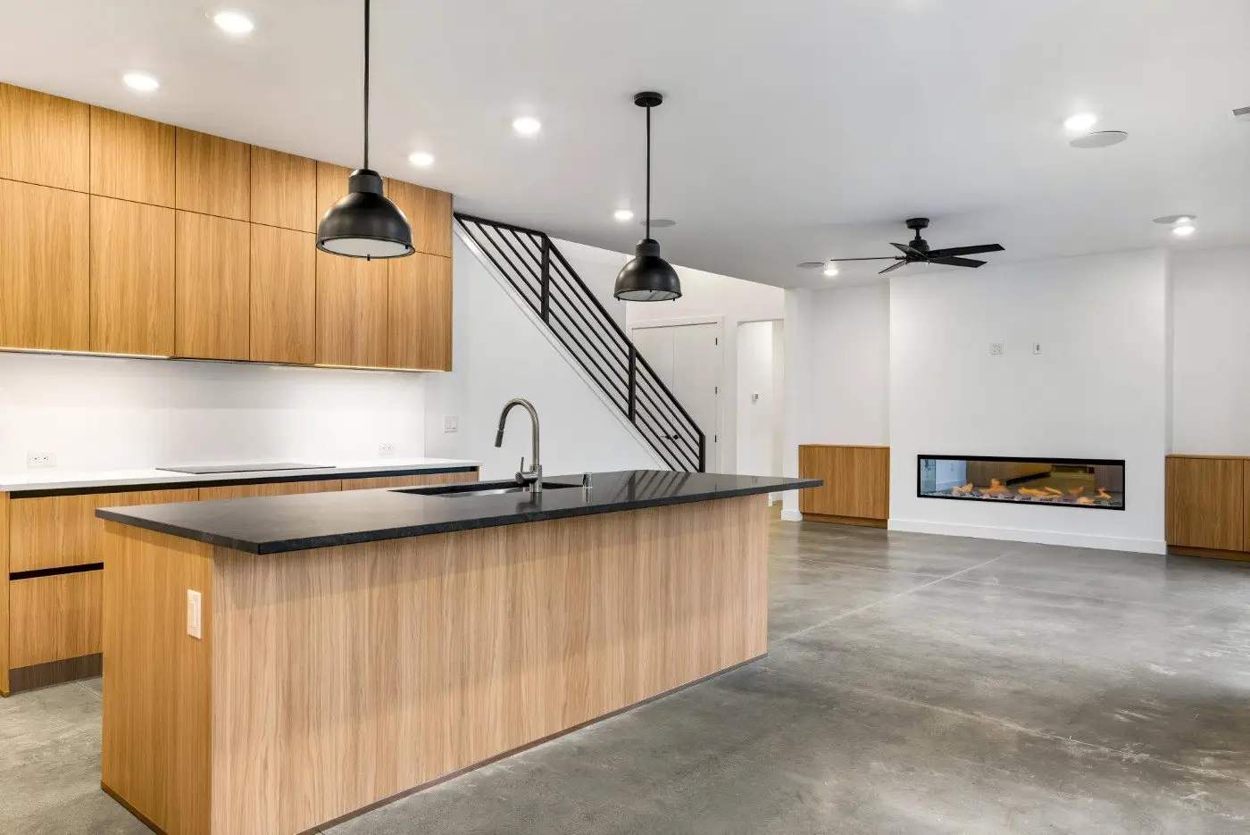 Modern kitchen with wood cabinets, a large island with a black countertop, black pendant lights, and concrete floors. In the background, there’s a staircase with a metal railing and a fireplace in the living area.