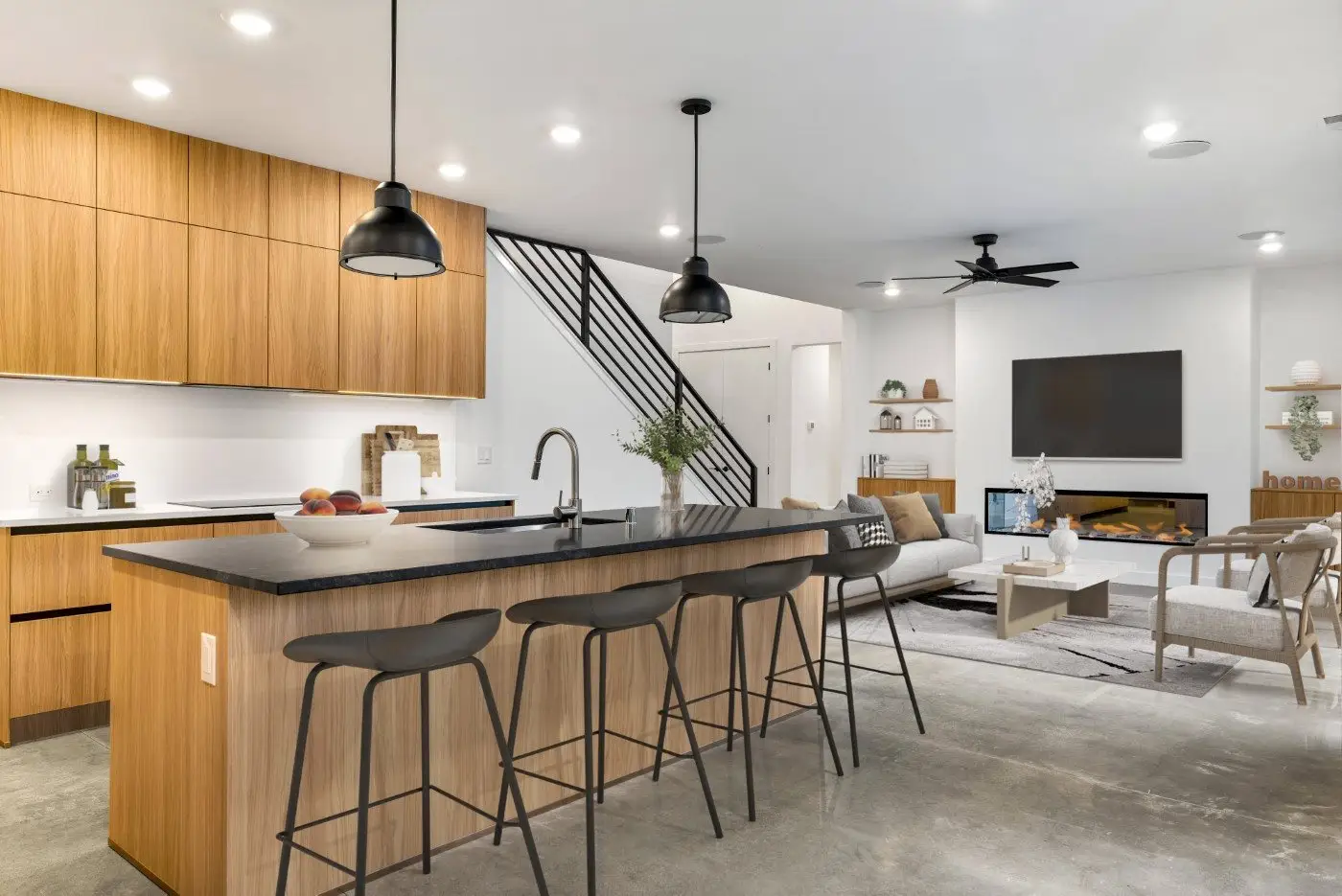 Modern open-concept kitchen and living room with wood cabinets, black countertops, bar stools, a cozy seating area, wall-mounted TV, fireplace, and minimalist decor. Exposed staircase and neutral tones throughout.